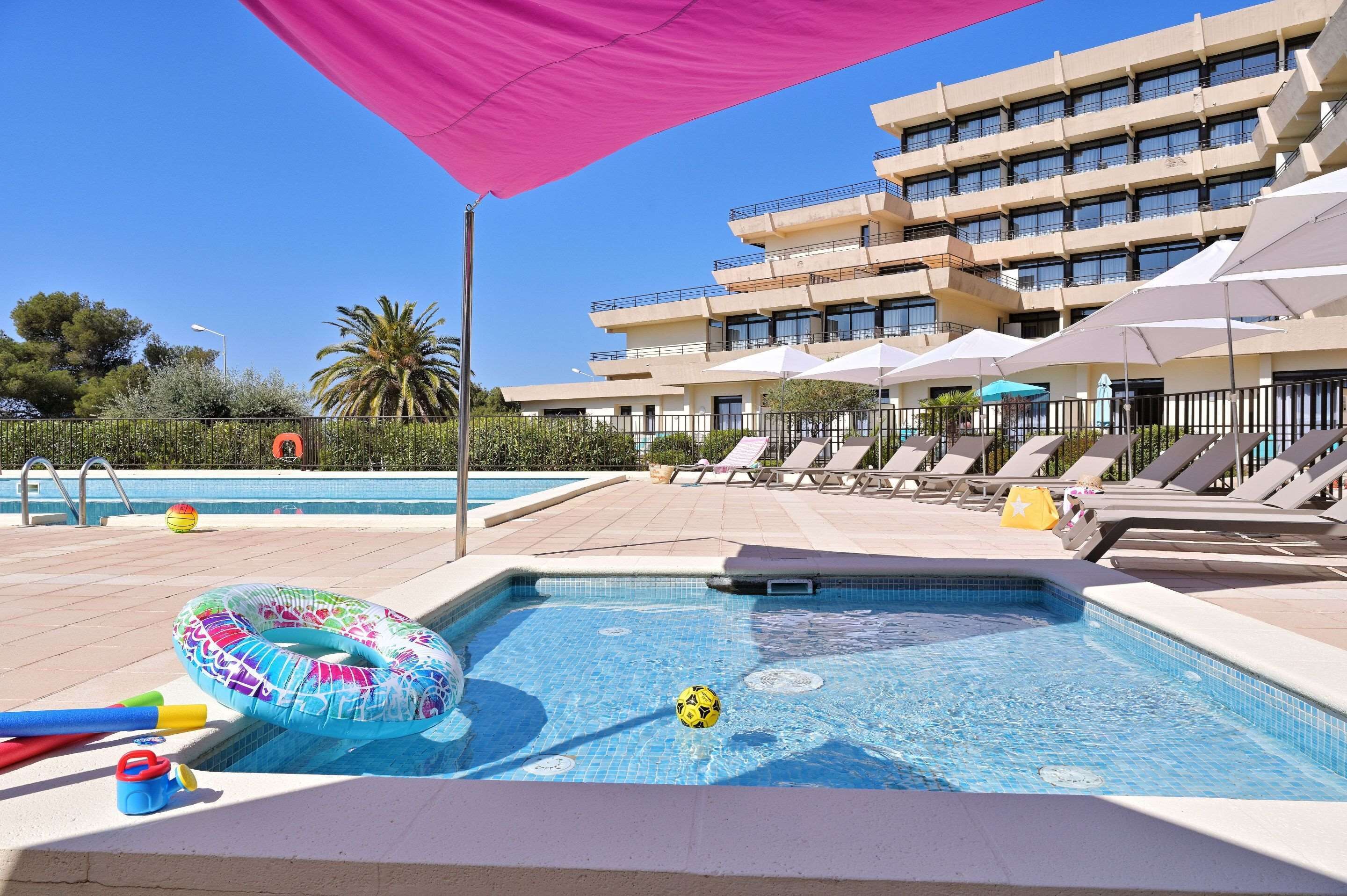Una piscina con sillas de descanso y sombrillas, rodeada por un edificio de varios pisos.