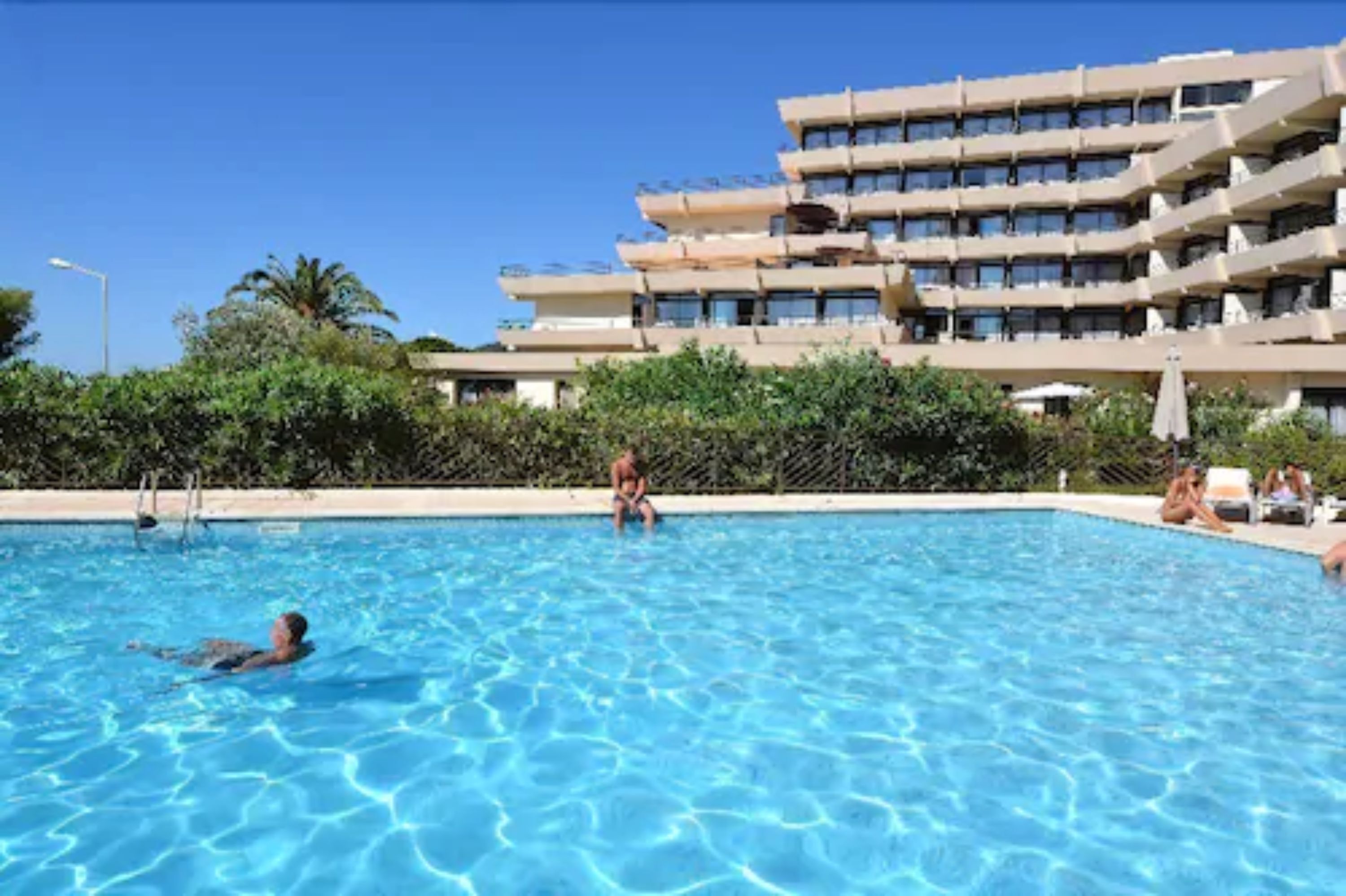 Une piscine entourée de personnes, un immeuble à plusieurs étages doté de balcons, et de la verdure en arrière-plan.