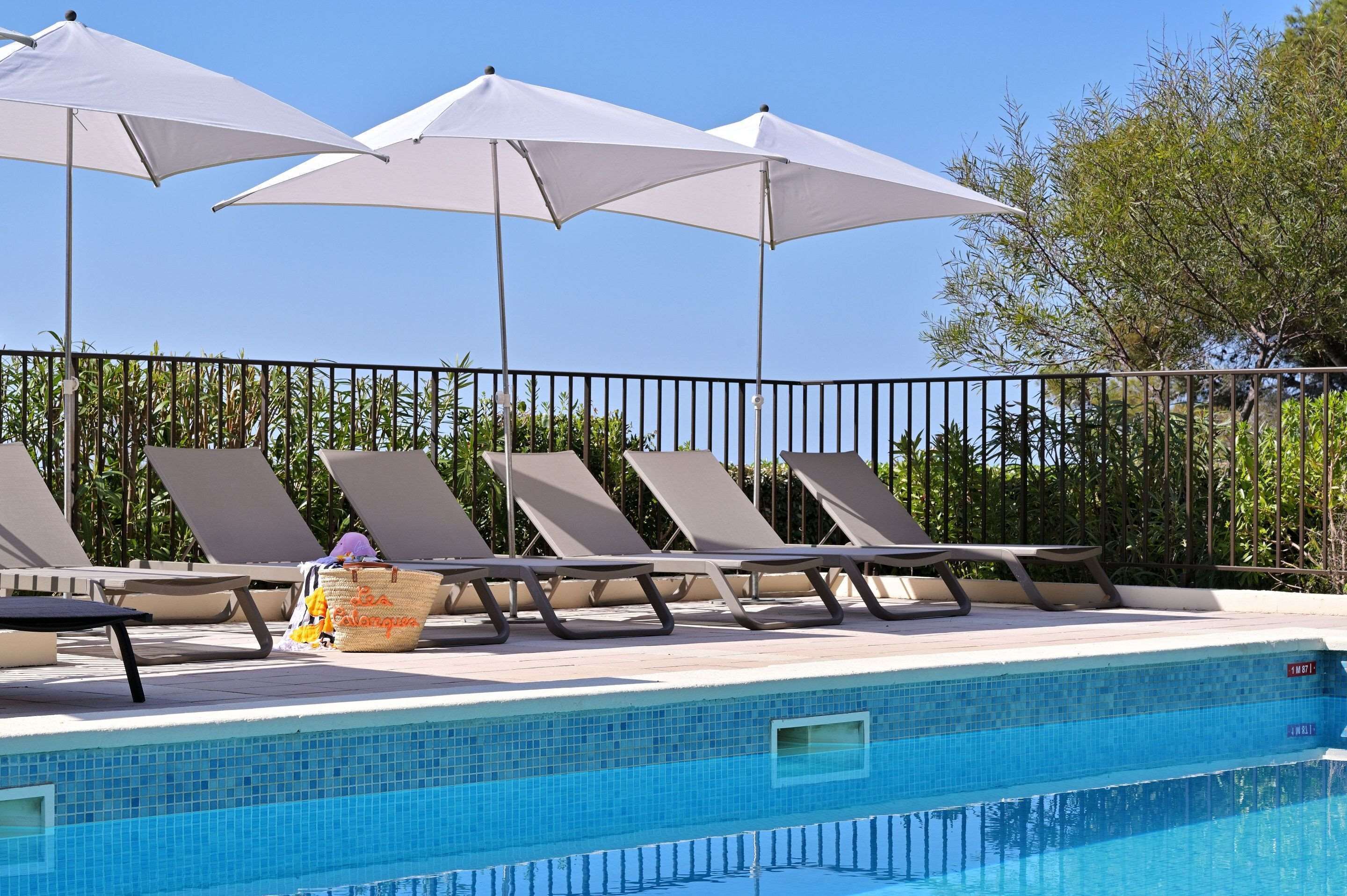 Zona de piscina amb gandules i ombrelles, una piscina amb rajoles i una tanca metàl·lica.
