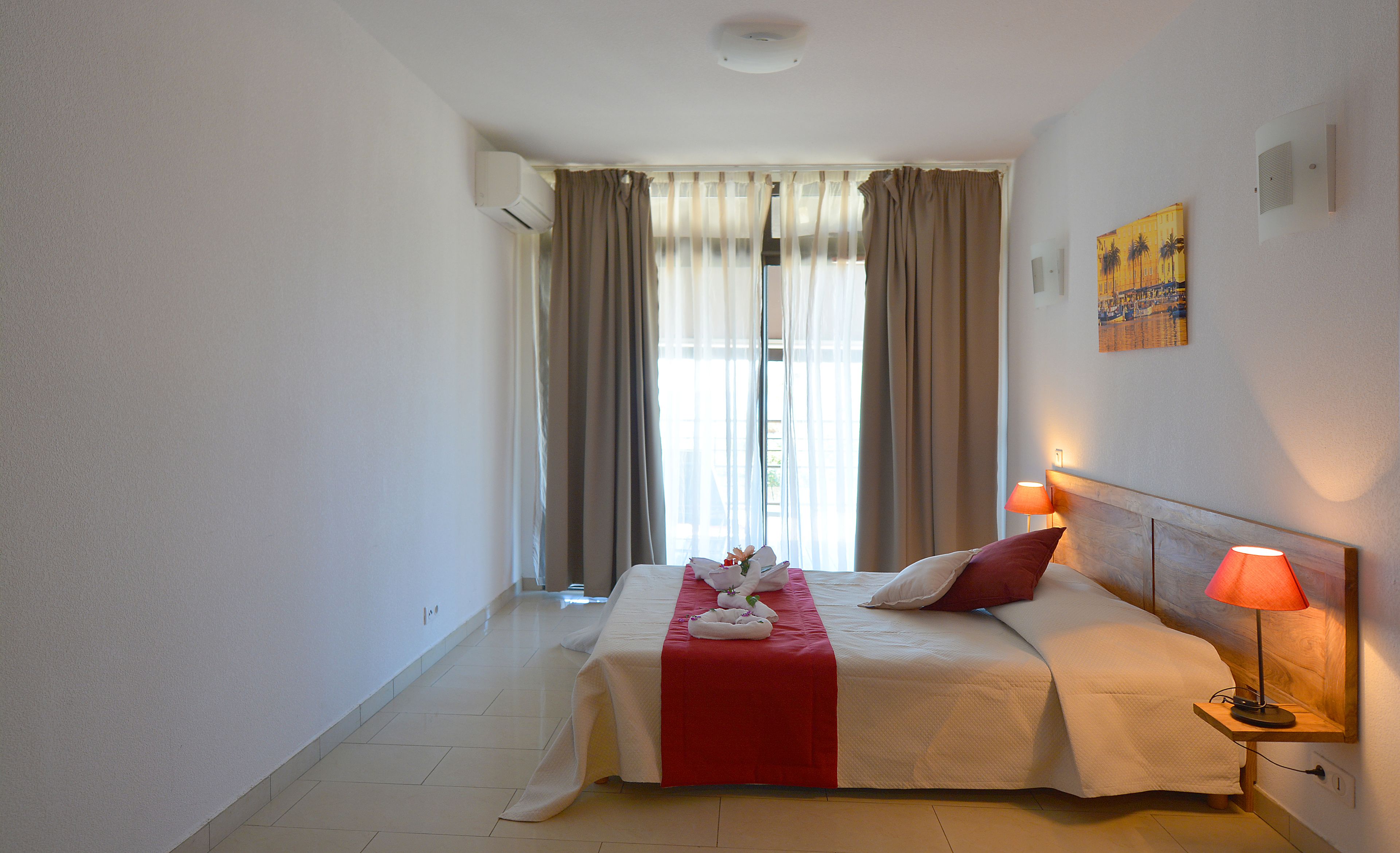 A hotel room with a bed, two bedside lamps, a window with curtains, and a wall-mounted air conditioner.