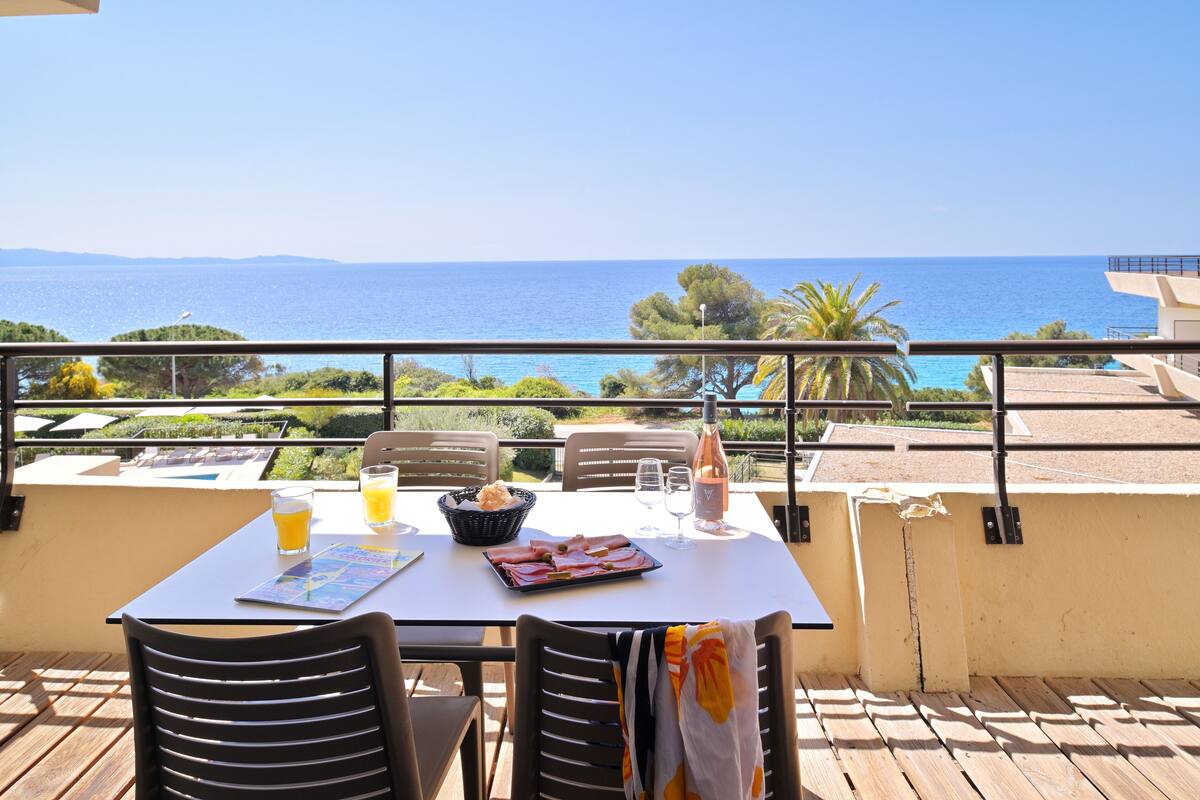 Una mesa con alimentos para el desayuno, una botella y vasos en un balcón con vistas al mar.