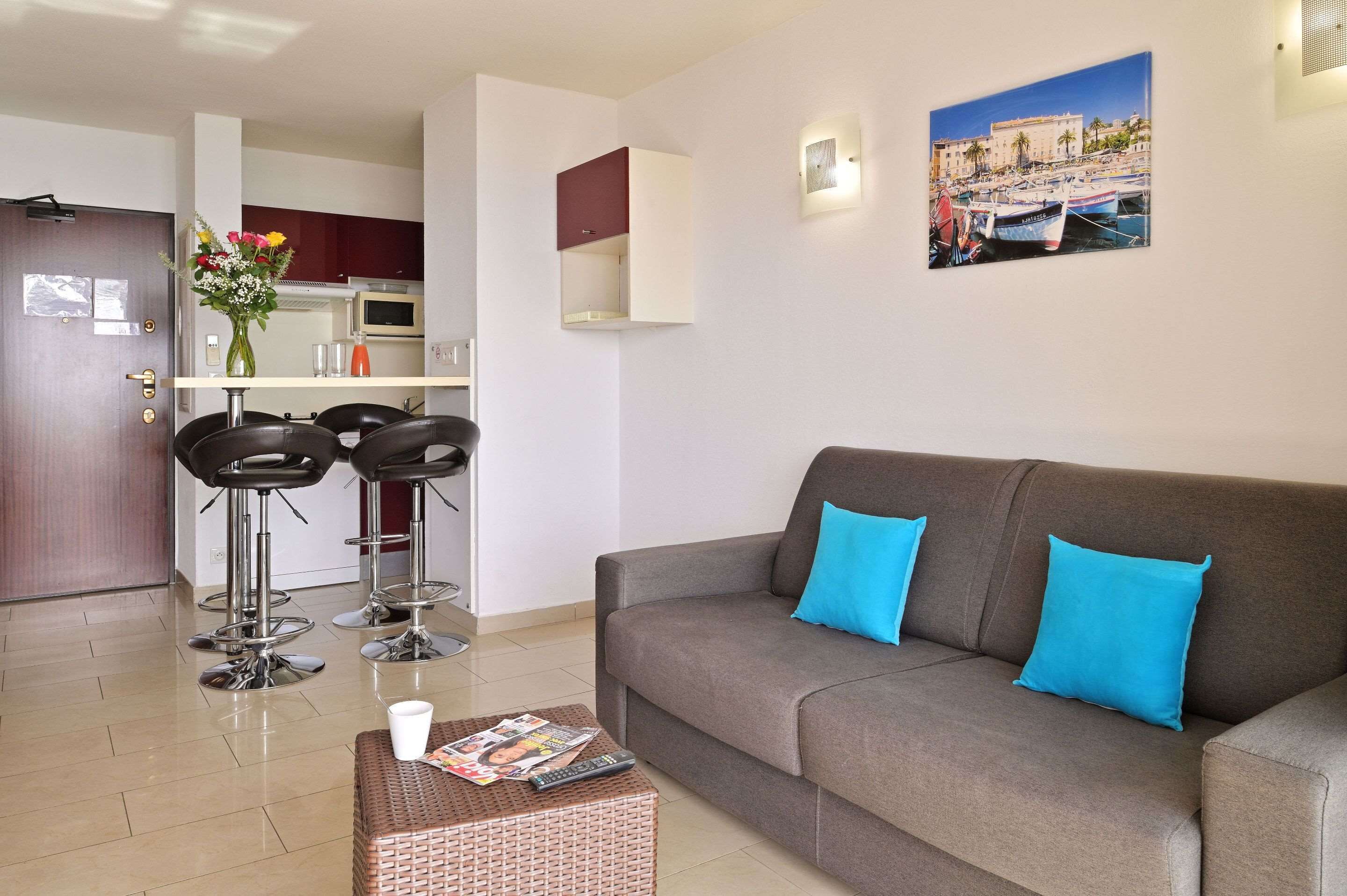 Ein modernes Wohnzimmer mit einem grauen Sofa, zwei blauen Kissen und einem Couchtisch mit Zeitschriften und einer Fernbedienung. Zwei schwarze Barhocker und eine Vase mit Blumen sind ebenfalls vorhanden. An der Wand hängt ein Gemälde einer Küstenlandschaft.