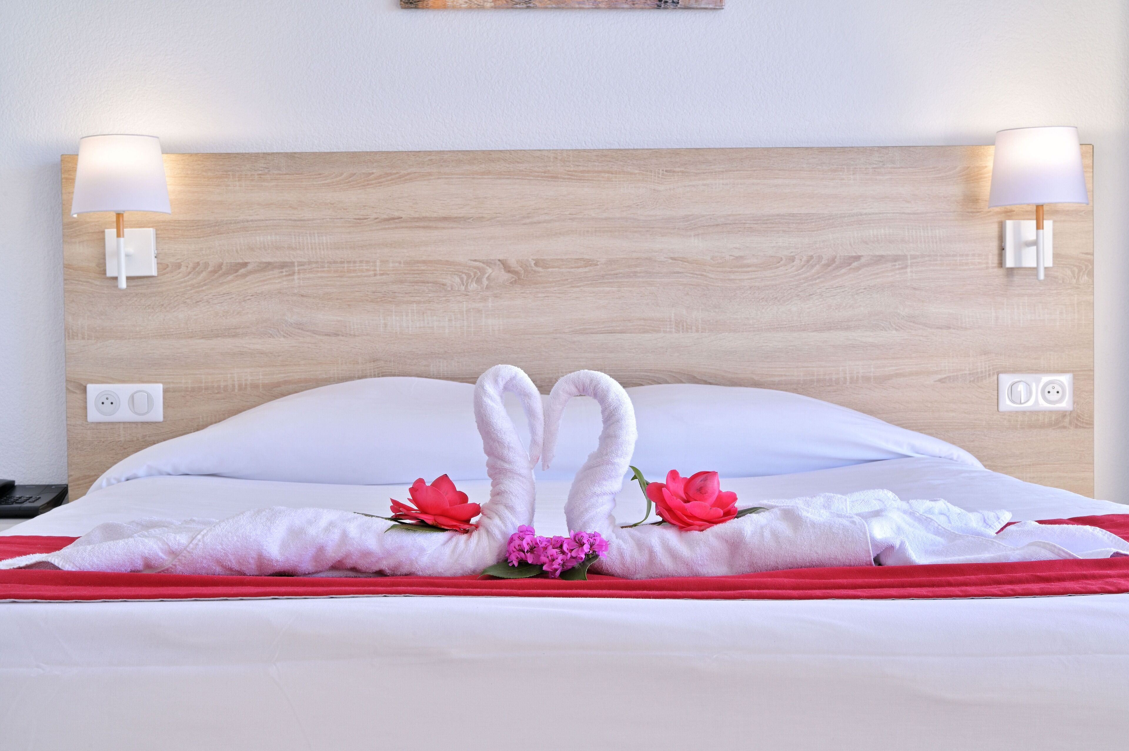 Una cama bien hecha con una colcha roja, dos toallas con forma de cisne y flores rojas sobre las almohadas.