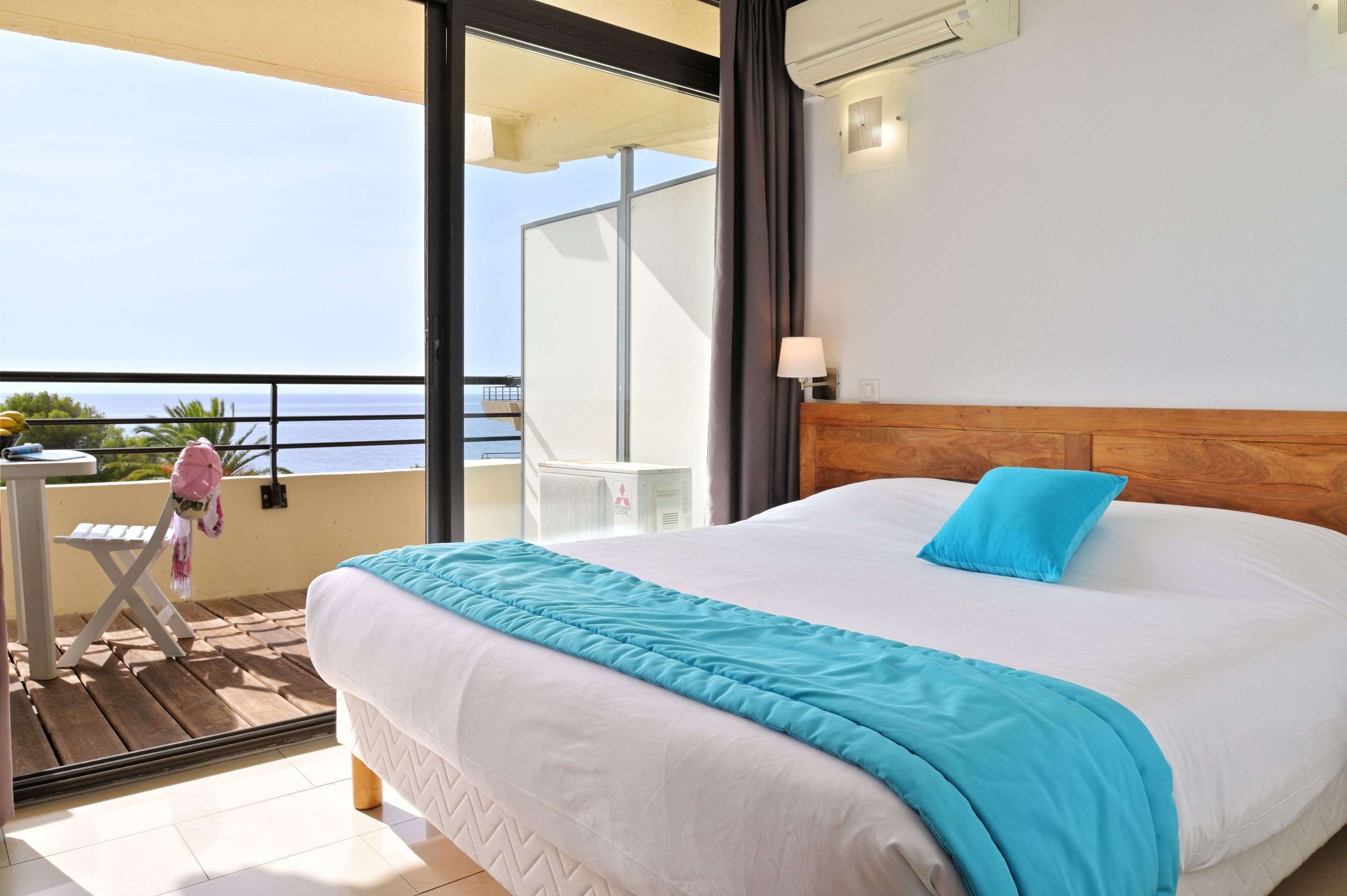A bedroom with a large bed, a balcony with a view of the sea, and a small table with a chair.