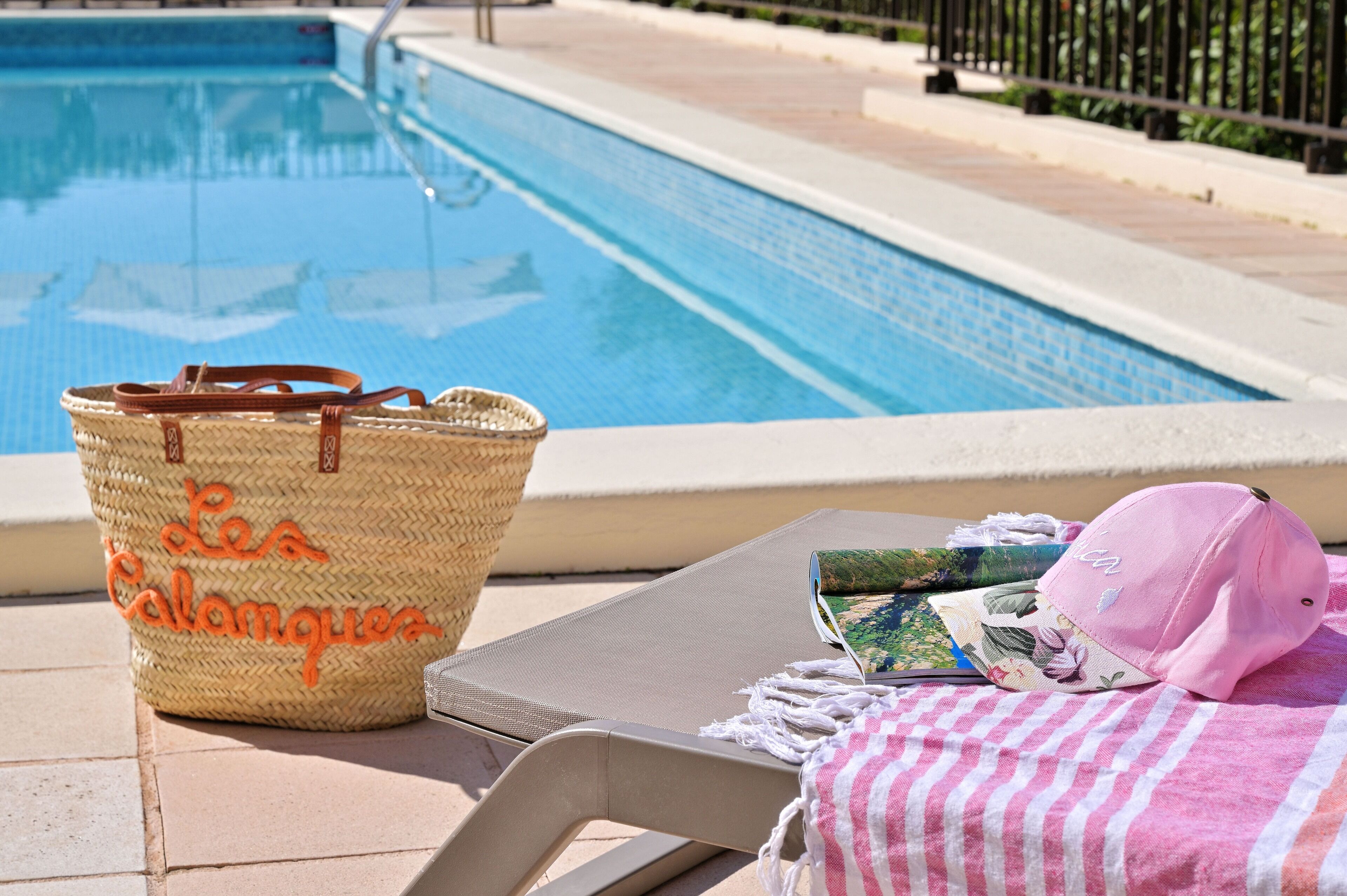 A poolside area with a woven basket, a beach chair, a pink cap, and a magazine on a towel.