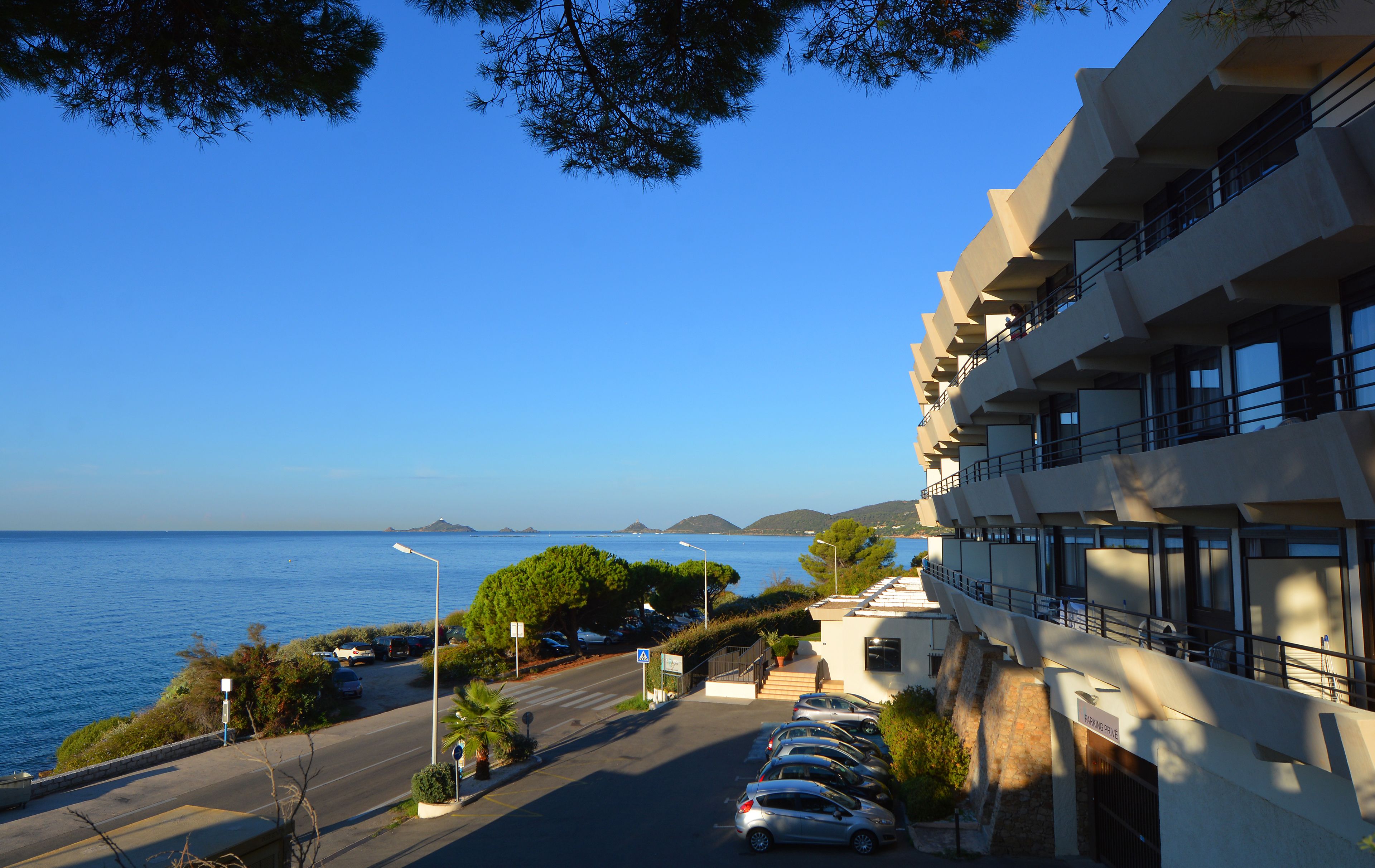 A coastal hotel with a view of the sea, parking, and a pedestrian pathway.