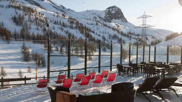 Paisaje nevado de una montaña con un área de comedor de restaurante que incluye mesas y sillas al aire libre.