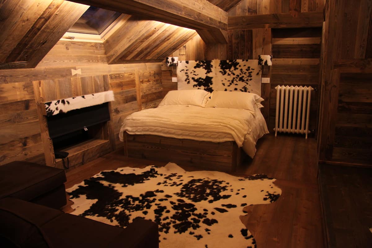 A rustic wooden room with a sloped ceiling, a bed with a cowhide rug, a fireplace, and a radiator.