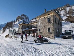 Ein Steinhaus mit Seilbahn und Schneemobil, umgeben von verschneitem Gelände und Bergen.