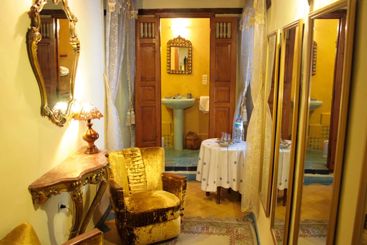 Un pasillo estrecho con un sillón antiguo, una mesa con una lámpara y un espejo a la izquierda. Hay una puerta que da a un baño con un lavamanos y una mesa redonda con mantel blanco.