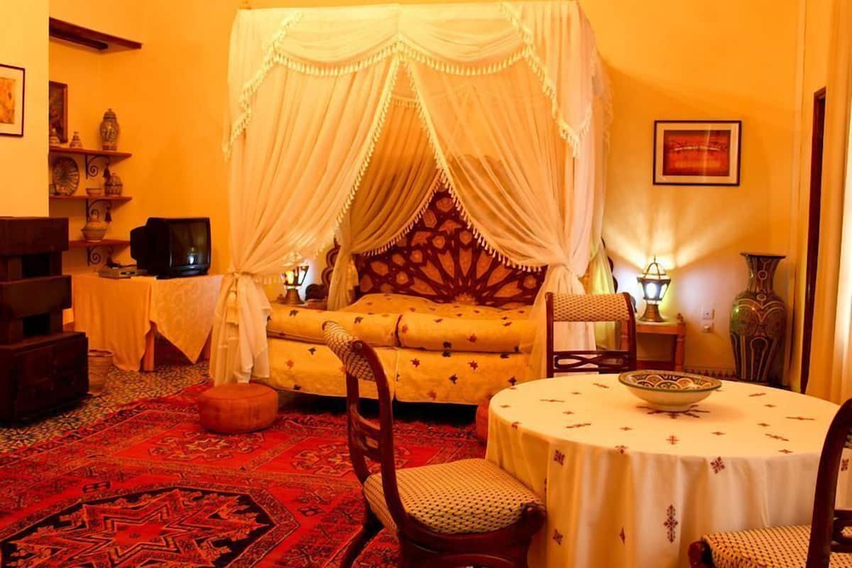Una habitación con una cama bajo un dosel, una mesa redonda con un cuenco, una silla, una mesita de noche con una lámpara y un televisor en un estante.