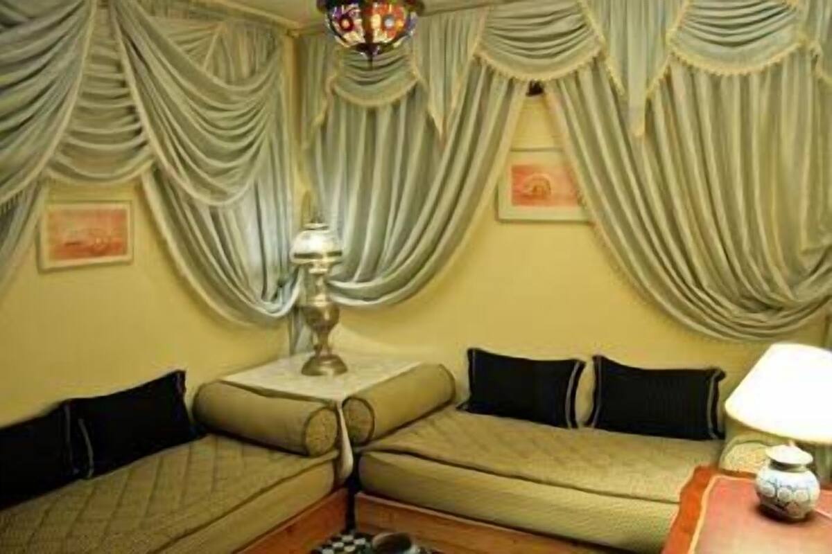 Una habitación con un sofá apoyado en una pared, una mesita con una lámpara y cortinas con drapeado.