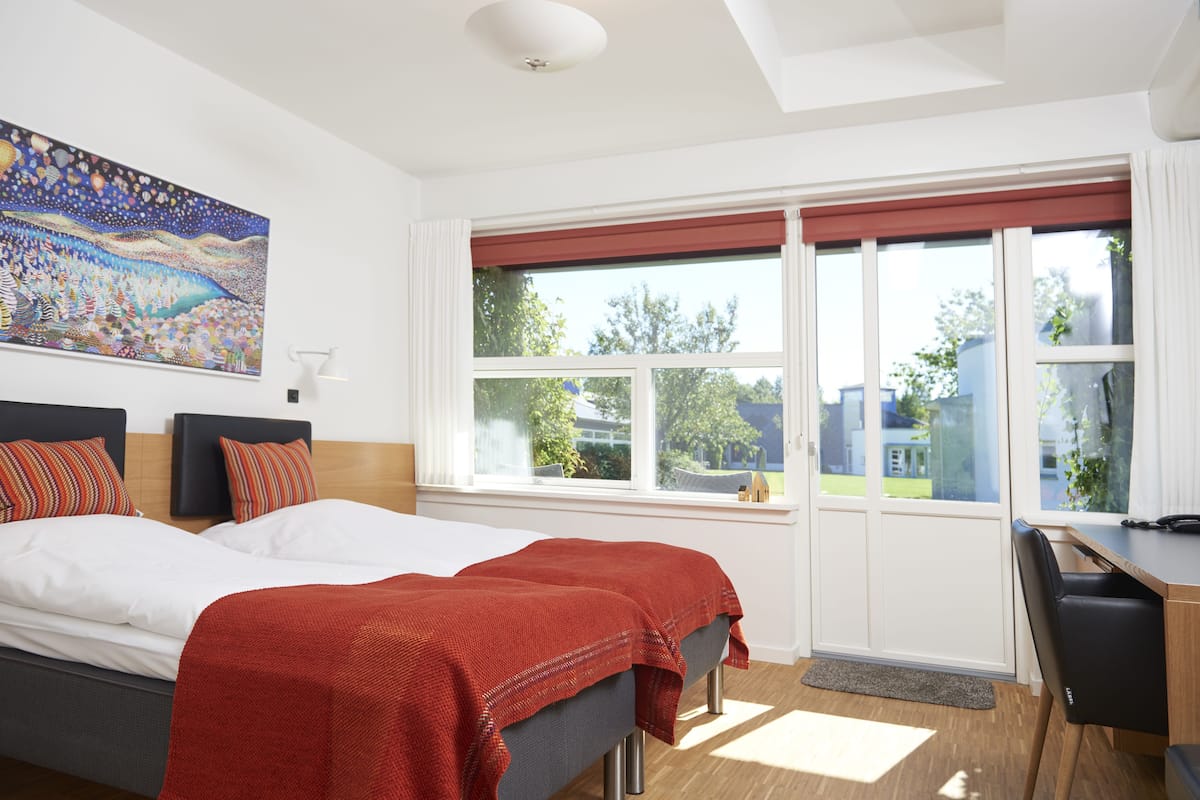 Habitación de hotel con dos camas, un escritorio y un ventanal con vistas al exterior.