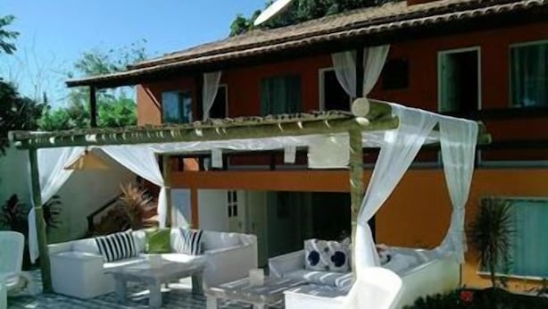 A covered outdoor seating area with white furniture, a patterned floor, and a building with a red facade and green shutters. - Pousada Elementos (Buzios)