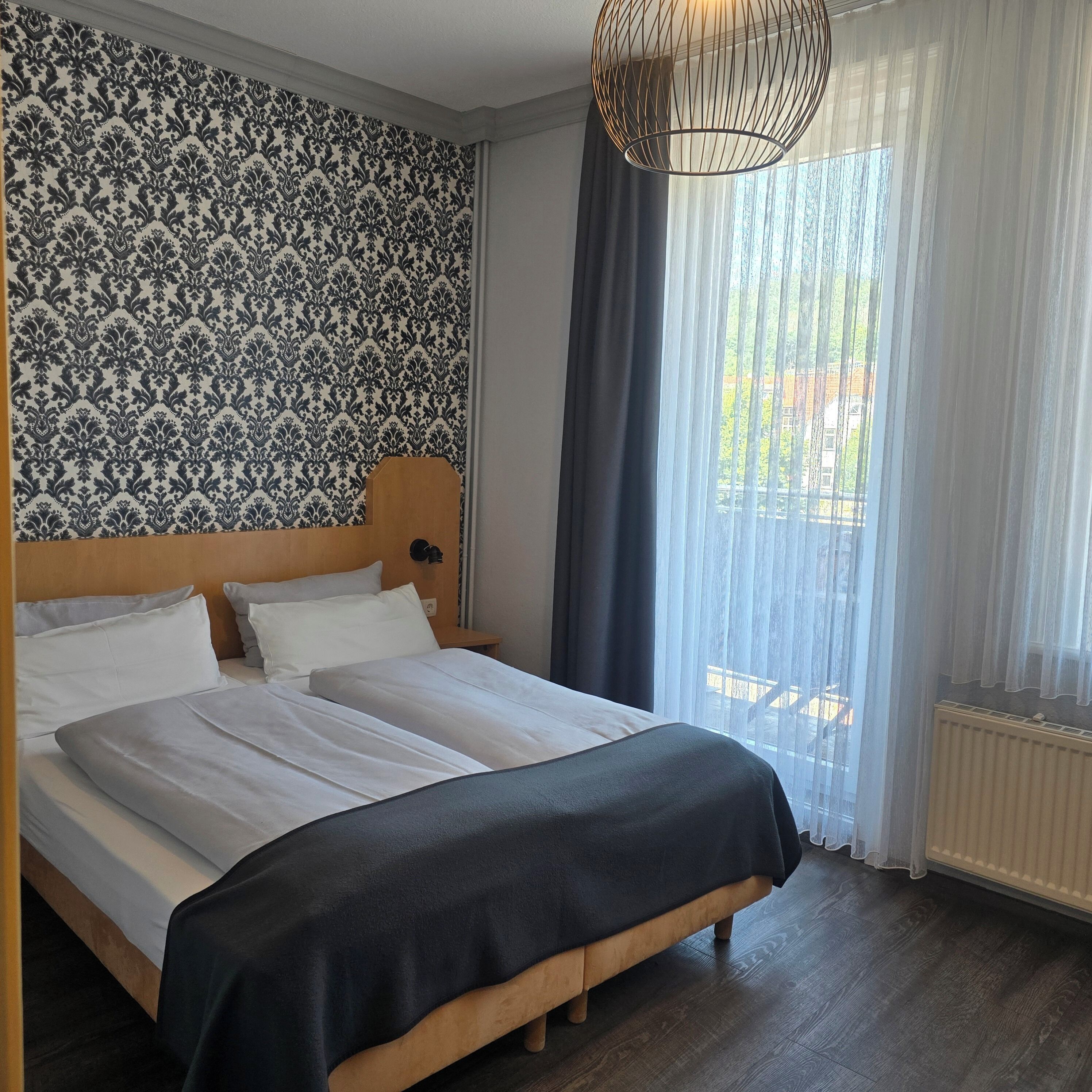 A double bed with white linens, a patterned wallpaper, a wooden headboard, and a large window with sheer curtains.