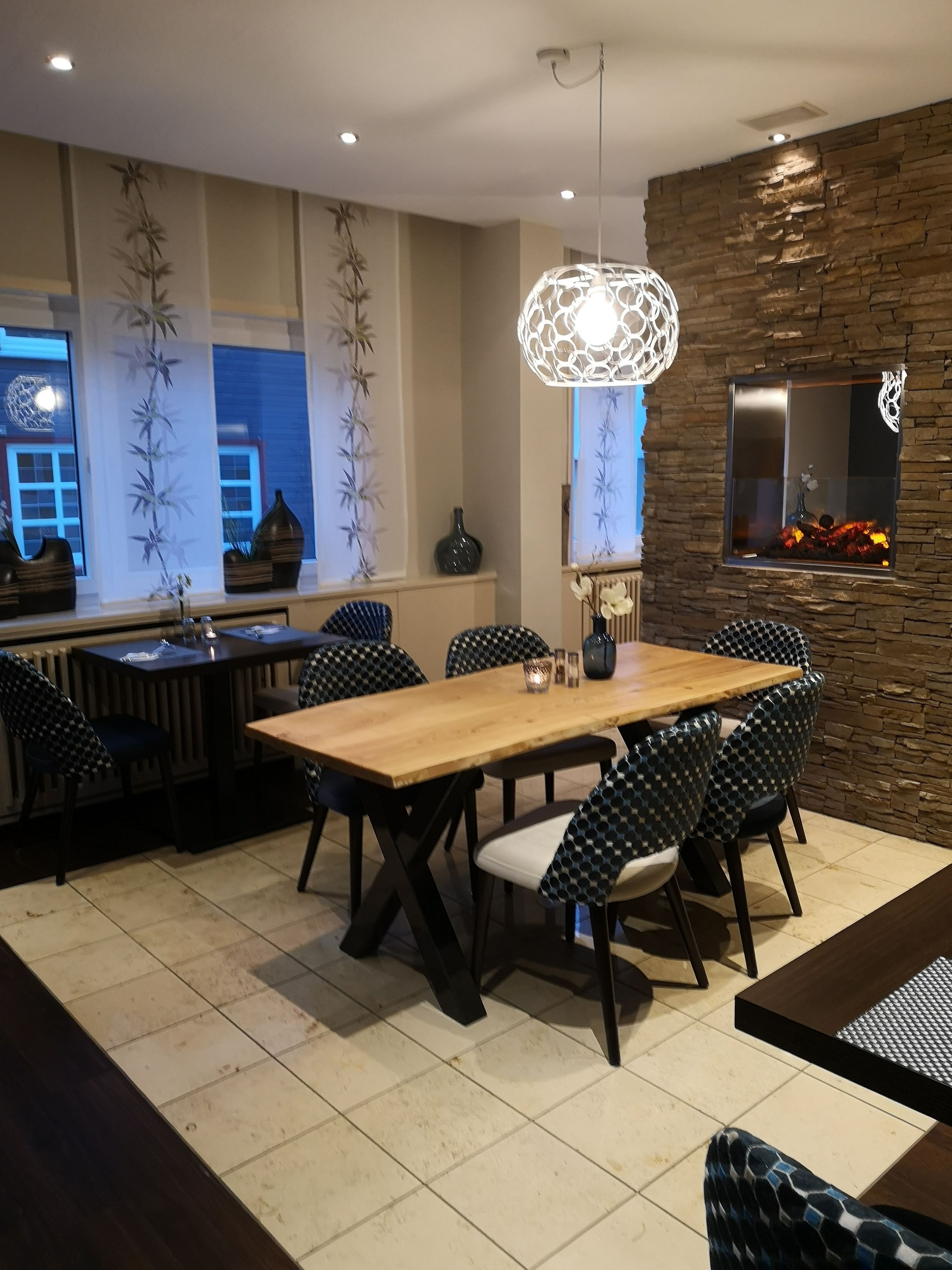 Uma sala de jantar moderna com uma mesa de madeira e cadeiras com padrão preto e branco. Há um lustre decorativo sobre a mesa e uma parede de pedra com lareira.