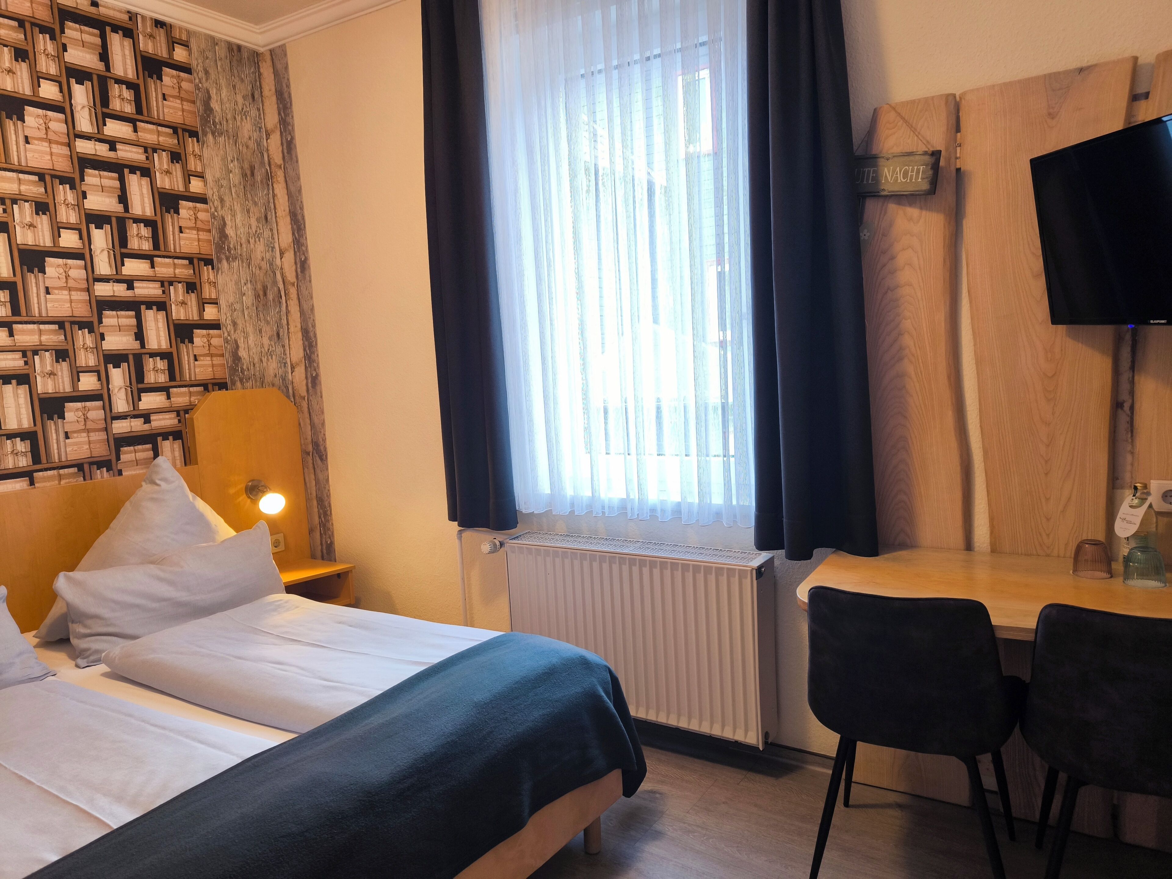 Ein Hotelzimmer mit zwei Betten, einem Schreibtisch, einem Sessel, einer Heizung, einem Fenster mit Vorhängen und einem an der Wand befestigten Fernseher.
