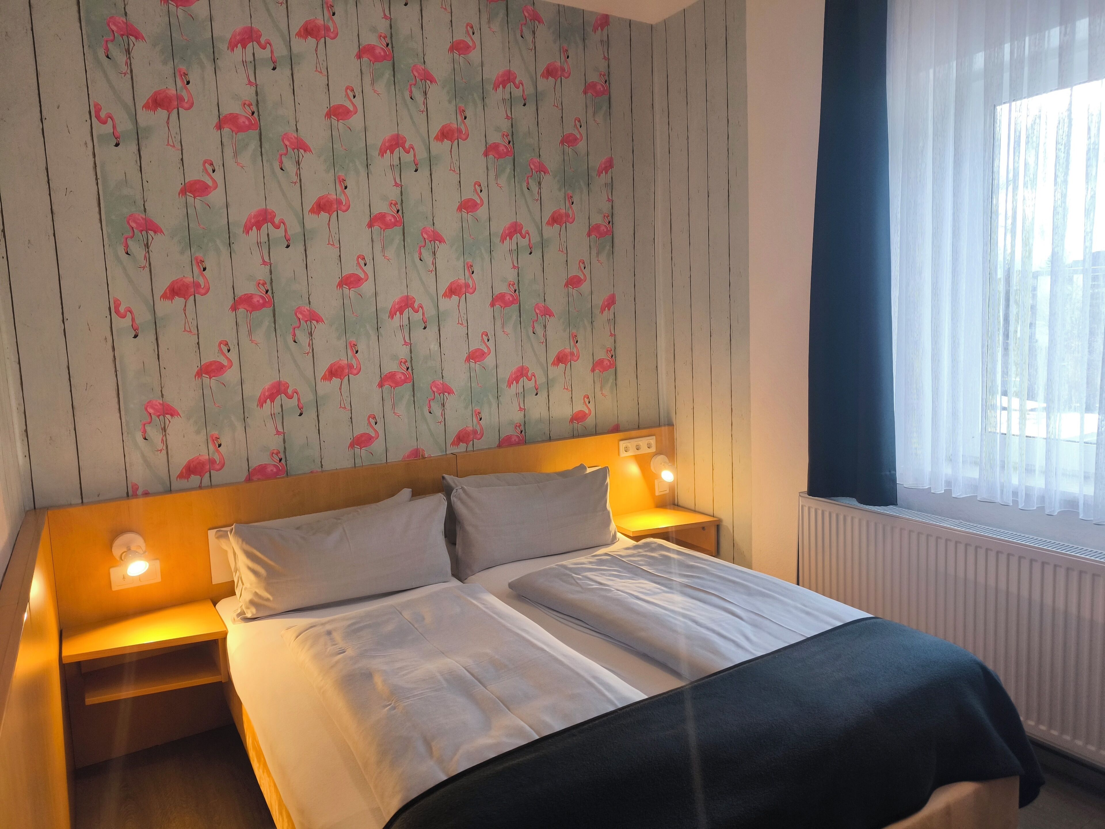A hotel room with a bed, two bedside tables, a nightstand, a window with sheer curtains, and a wall with a flamingo wallpaper pattern.