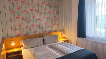 A hotel room with a bed, two bedside tables, a nightstand, a window with sheer curtains, and a wall with a flamingo wallpaper pattern.