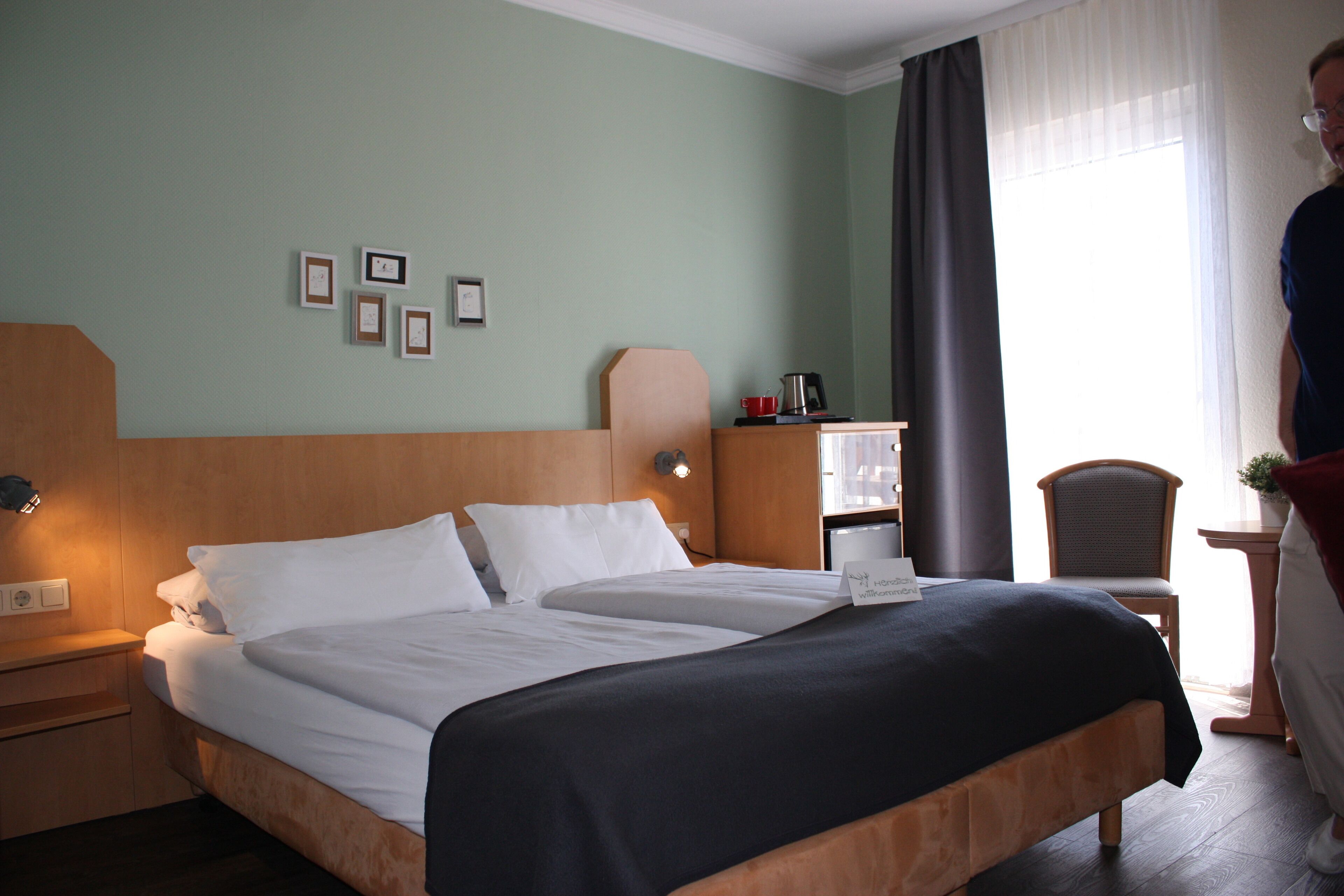 Een hotelkamer met een groot bed, een nachtkastje met een lamp, een klein bureau met een stoel, en een raam met gordijnen.