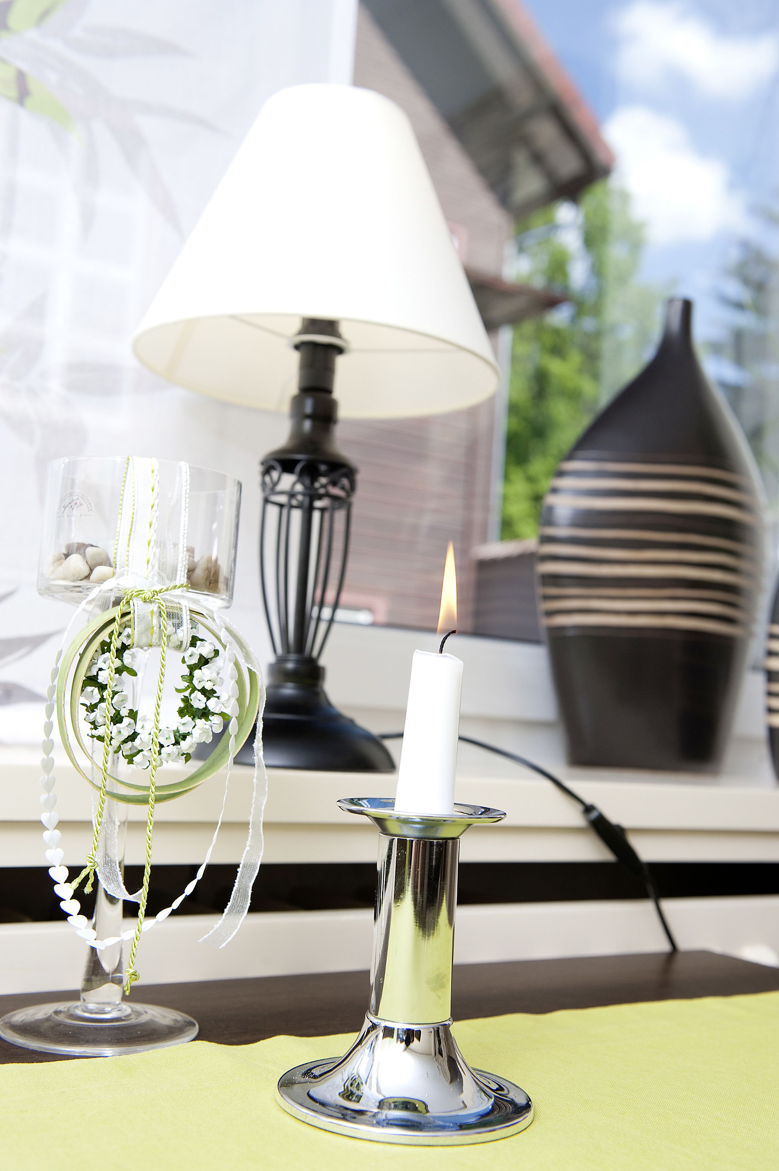 Una mesa con una vela encendida, un candelabro de vidrio con una guirnalda de cuentas blancas y un vaso decorado con flores blancas.