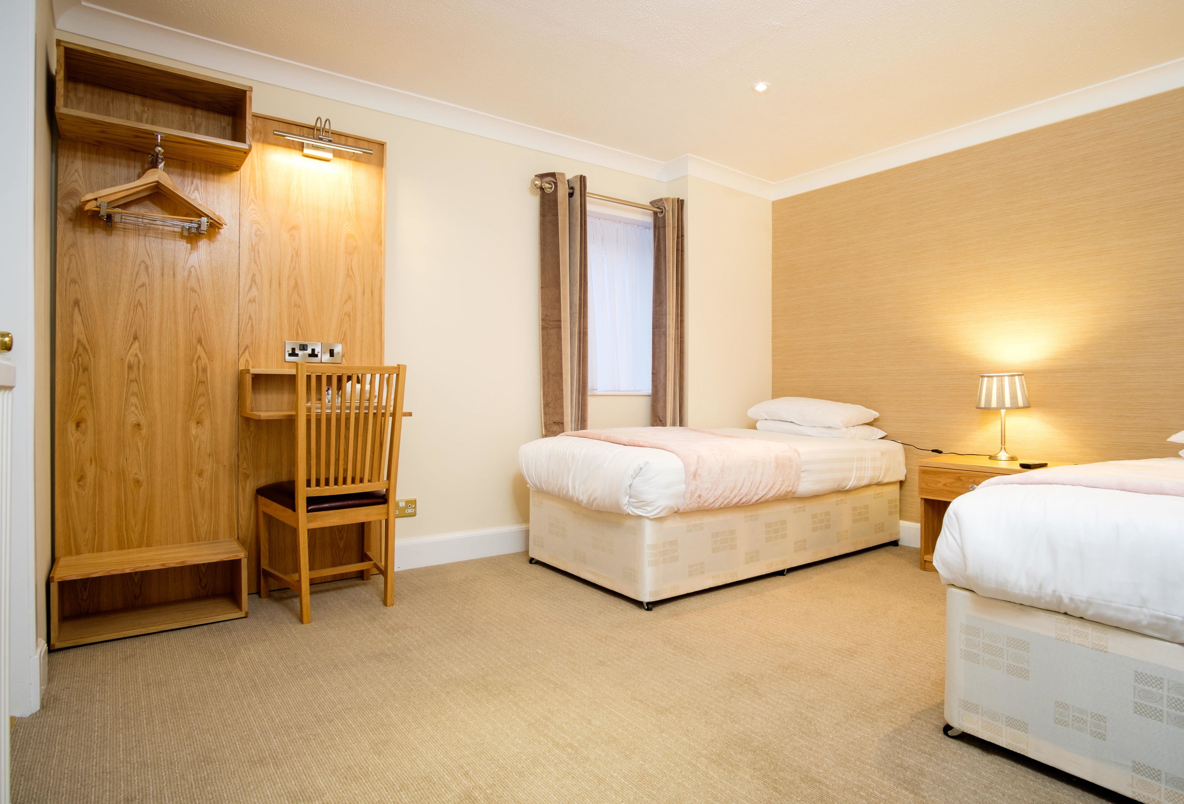 A hotel room with two beds, a wooden wardrobe, a chair, a bedside table with a lamp, and a window with curtains.