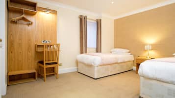 A hotel room with two beds, a wooden wardrobe, a chair, a bedside table with a lamp, and a window with curtains.