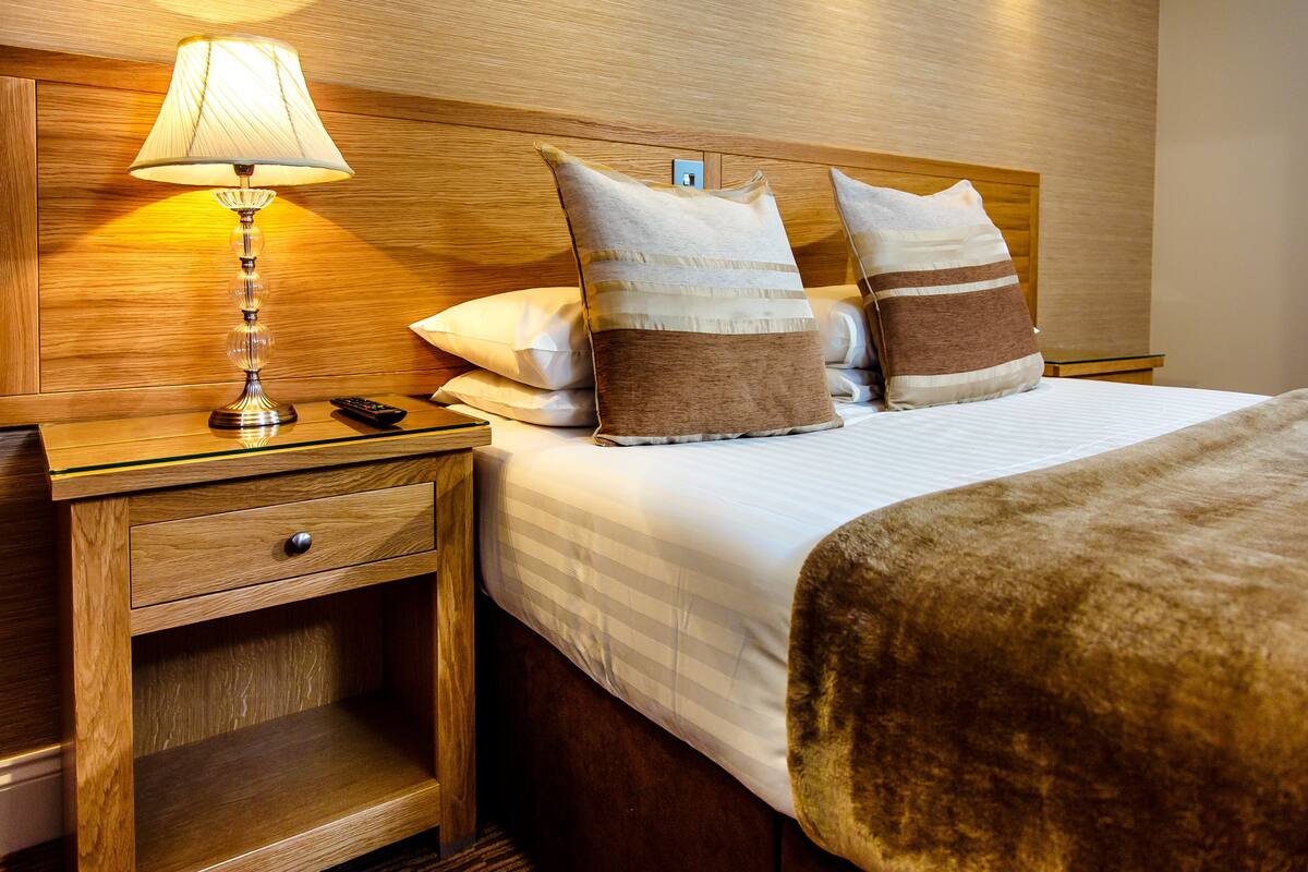 Habitación de hotel impecable, con cabecera de madera, lámpara de noche, mesita de noche con cajón y dos almohadas.
