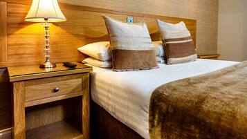 Habitación de hotel impecable, con cabecera de madera, lámpara de noche, mesita de noche con cajón y dos almohadas.