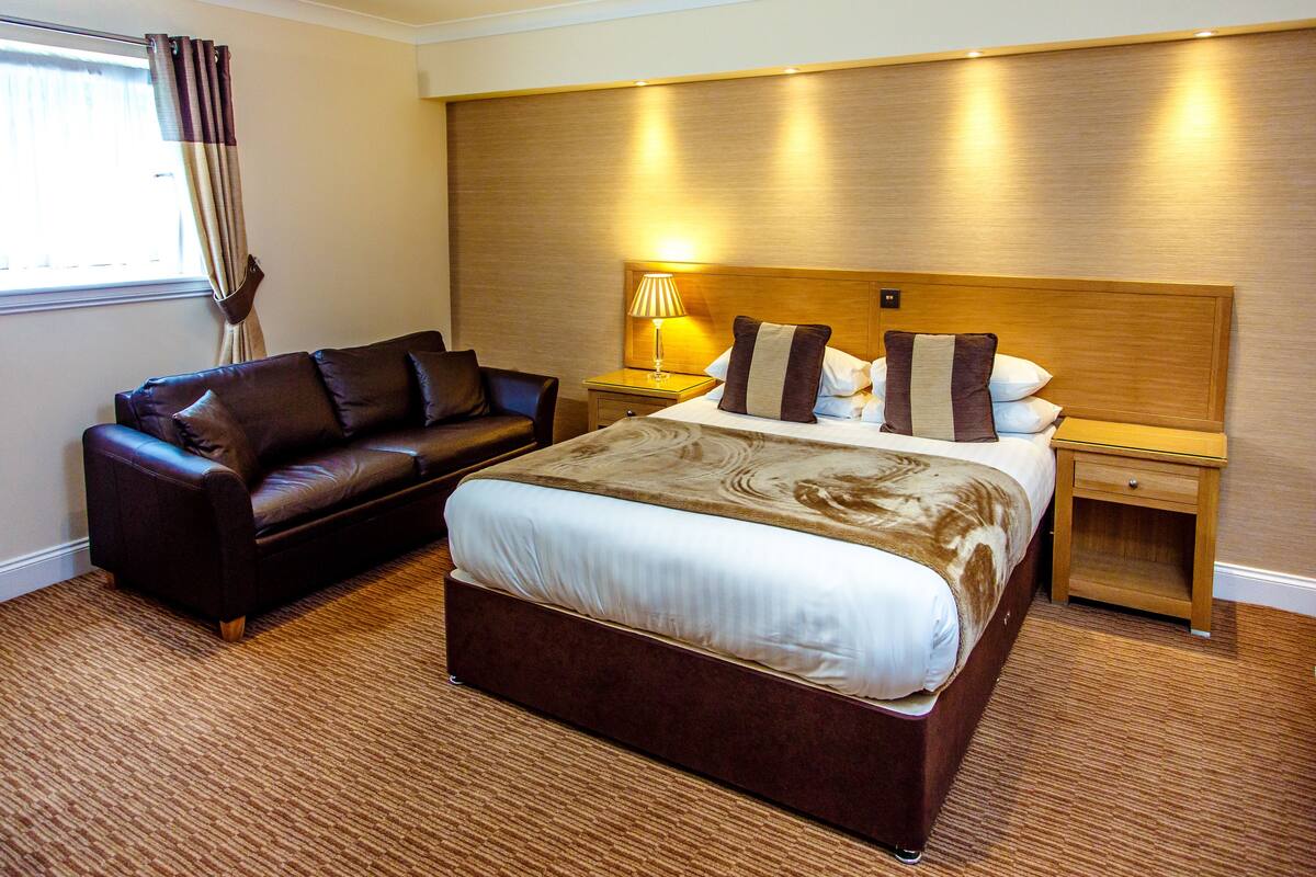 Una habitación de hotel con una cama grande, un sofá de cuero, una mesita de noche con lámpara y un cabecero de madera.