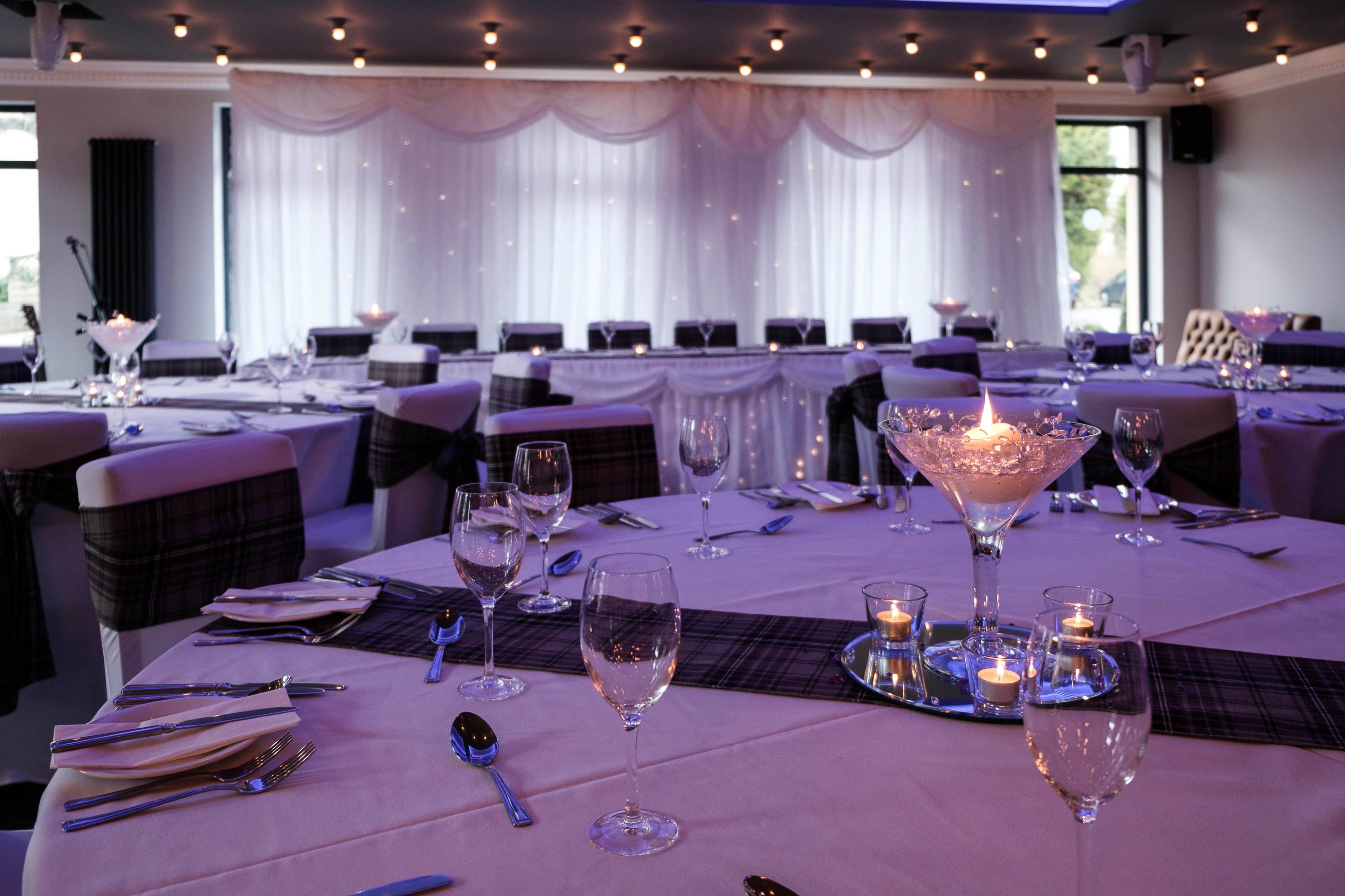 A dining area with tables set for a formal event, featuring candles, wine glasses, and cutlery.