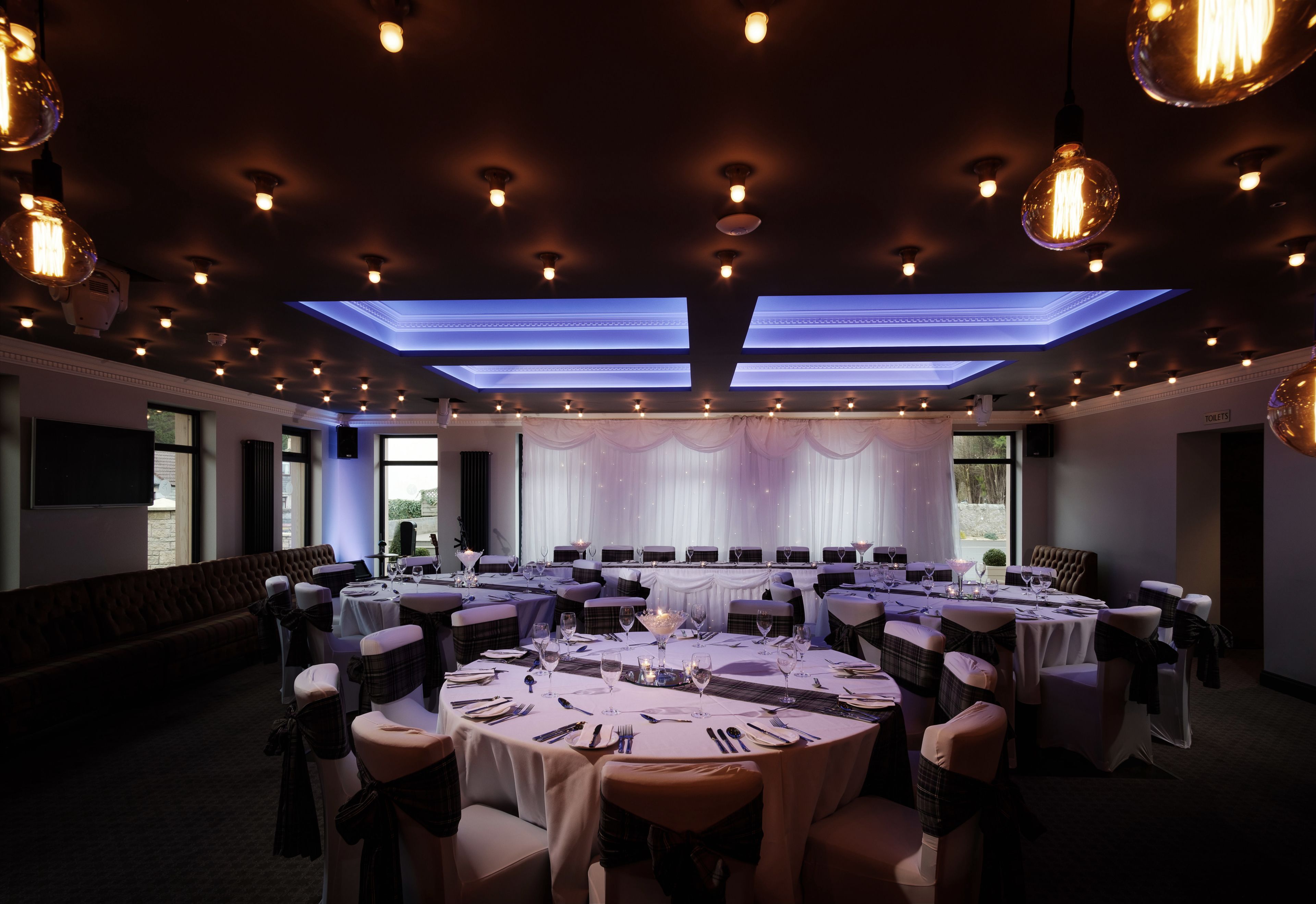 A banquet hall with round tables set for dining, a stage with curtains, and a seating area with a sofa.