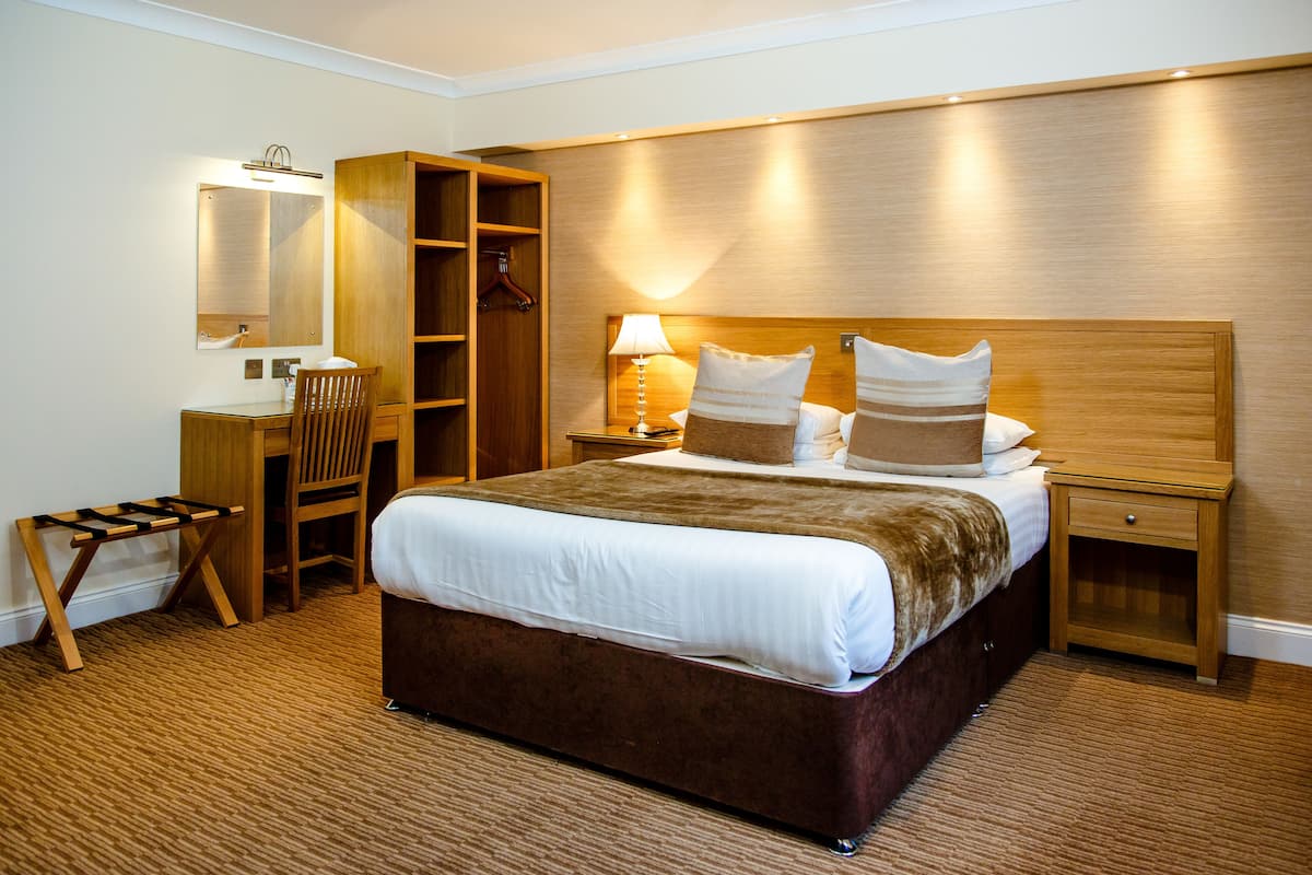 Habitación de hotel con una cama grande, un escritorio con silla, un armario y una mesita de noche con lámpara.