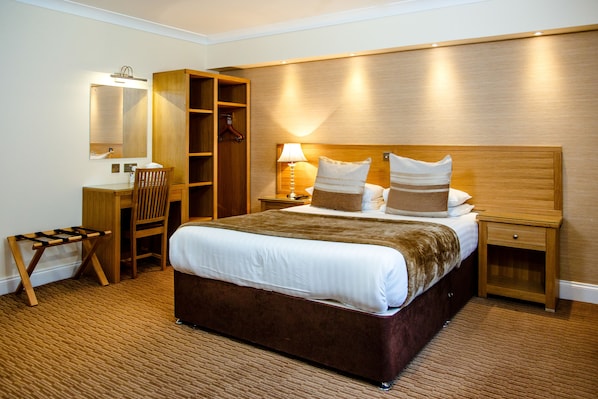 A hotel room with a large bed, a desk with a chair, a wardrobe, and a bedside table with a lamp.