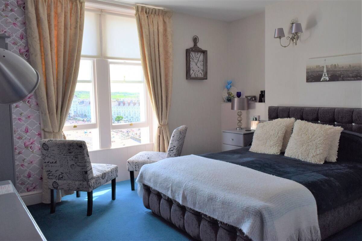 A bedroom with a large bed, a chair, a nightstand, a lamp, a clock, and a window with curtains.
