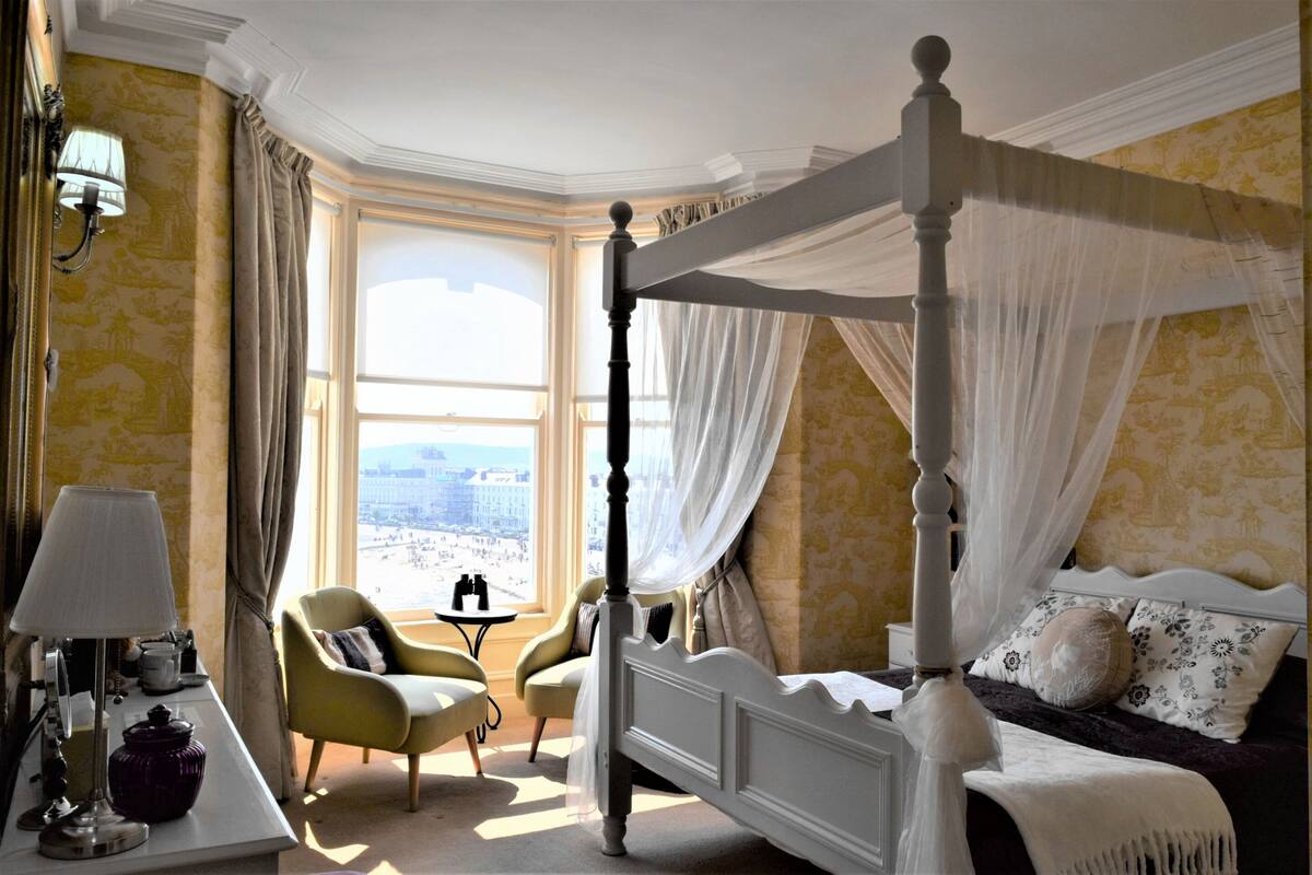 A four-poster bed with sheer curtains, a green armchair, a small table with a lamp, and a view of the city through large windows.
