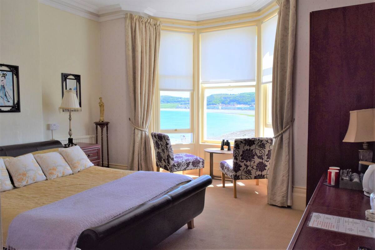 A hotel room with a large bed, two chairs, a small table, and a view of the sea through a bay window.