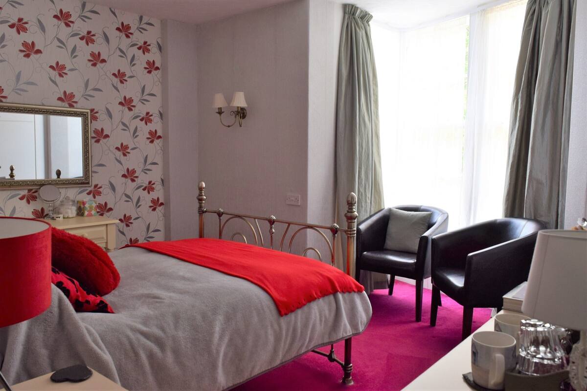 A hotel room with a bed, two chairs, a desk, a mirror, and a window with curtains.