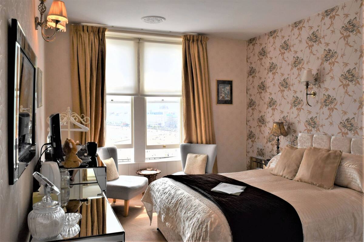 A hotel room with a bed, a desk with a chair, a mirror, and a window with curtains.