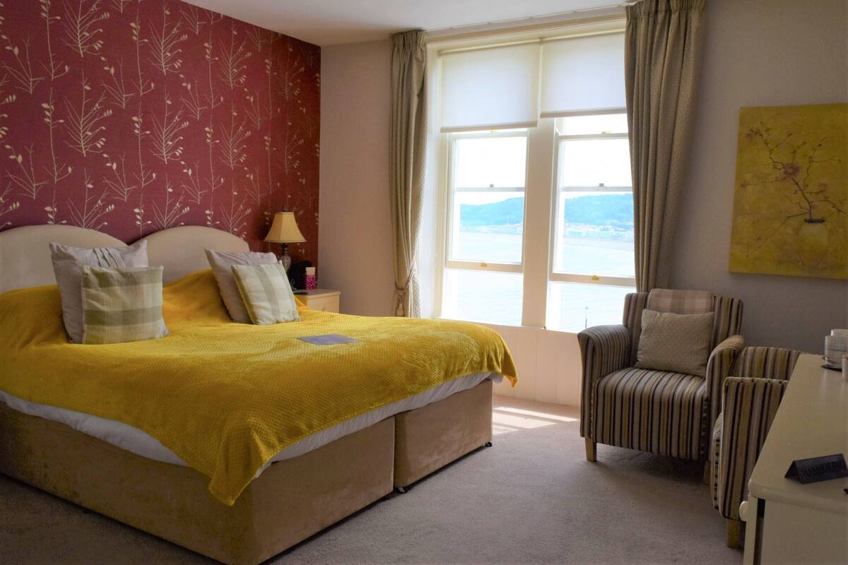 A bedroom with a bed, a chair, a nightstand, a lamp, and a window with a view.