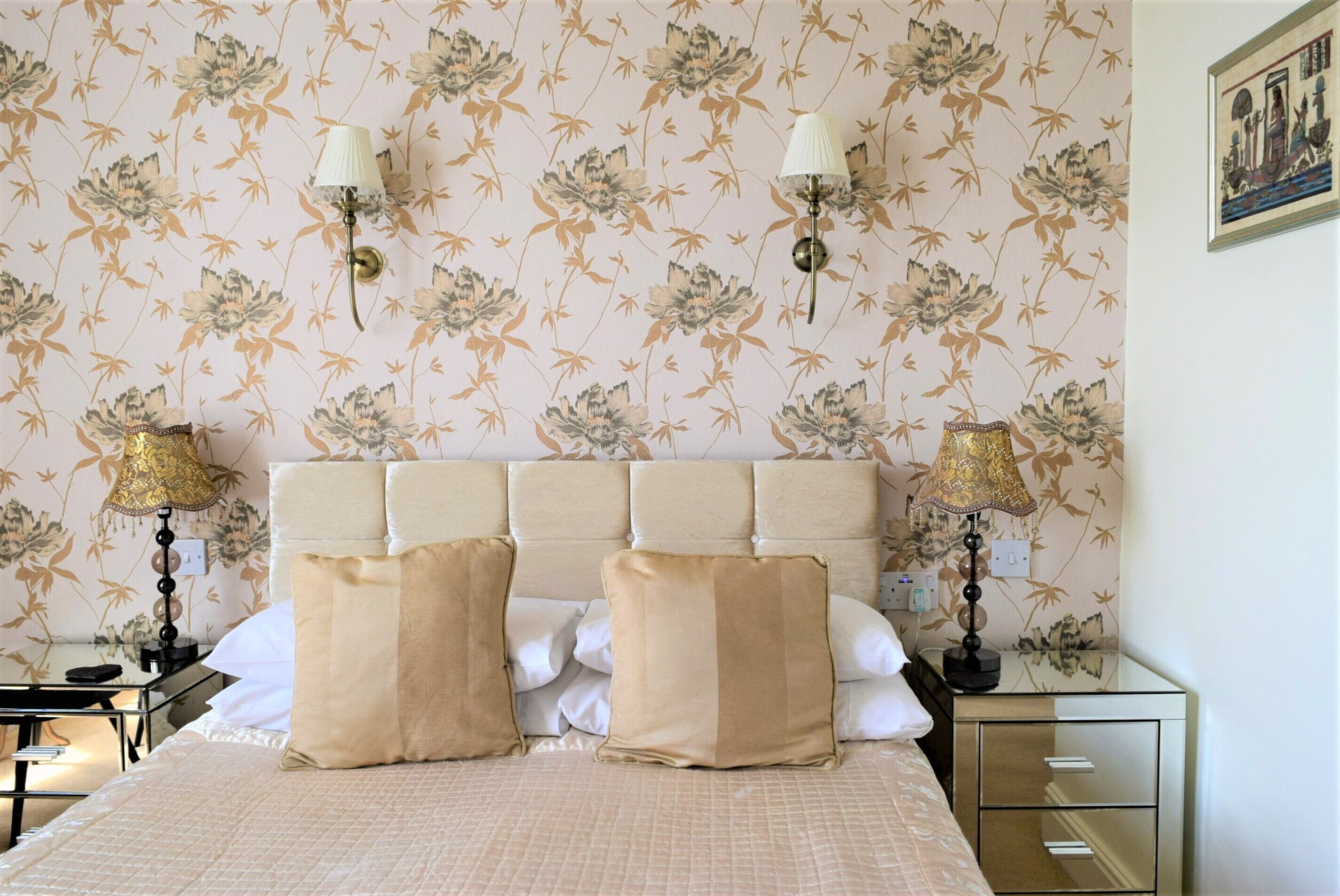 Una habitación de hotel con papel tapiz floral, un cabecero capitonado, dos lámparas de noche, una cama con ropa de cama color beige y una mesita de noche con lámpara.