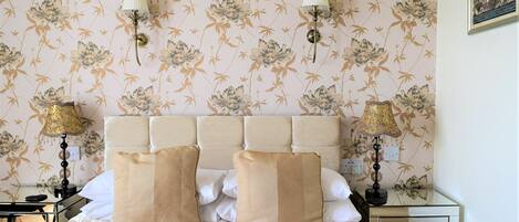 Una habitación de hotel con papel tapiz floral, un cabecero capitonado, dos lámparas de noche, una cama con ropa de cama color beige y una mesita de noche con lámpara.