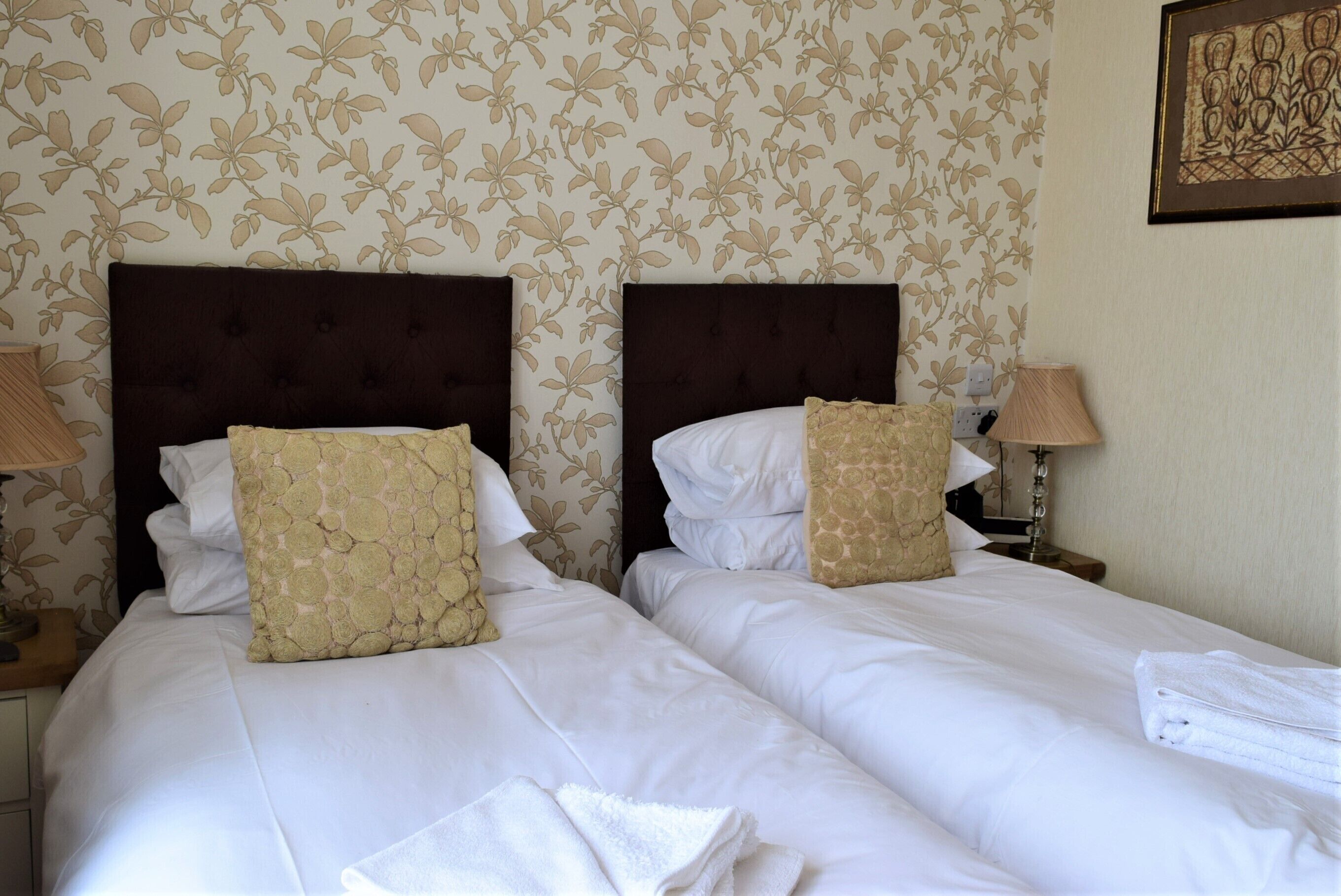 A hotel room with two beds, each with a dark headboard and white bedding. There are two bedside tables with lamps, and a wall-mounted artwork above the beds.