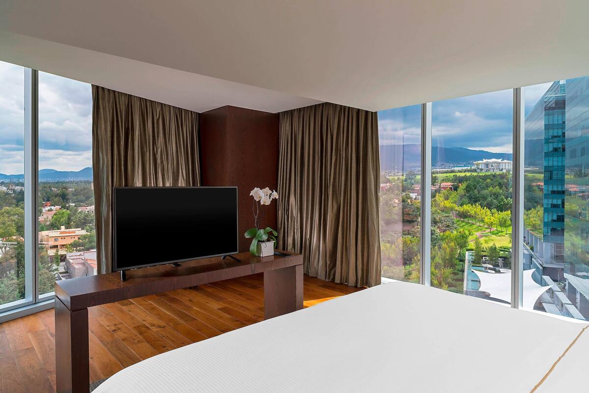 Una moderna camera d'albergo con una televisione di grandi dimensioni su un supporto in legno, un letto e vista su una città e montagne attraverso ampie finestre.