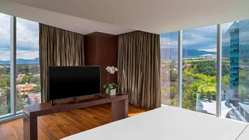 A modern hotel room with a large TV on a wooden stand, a bed, and a view of a cityscape and mountains through large windows.