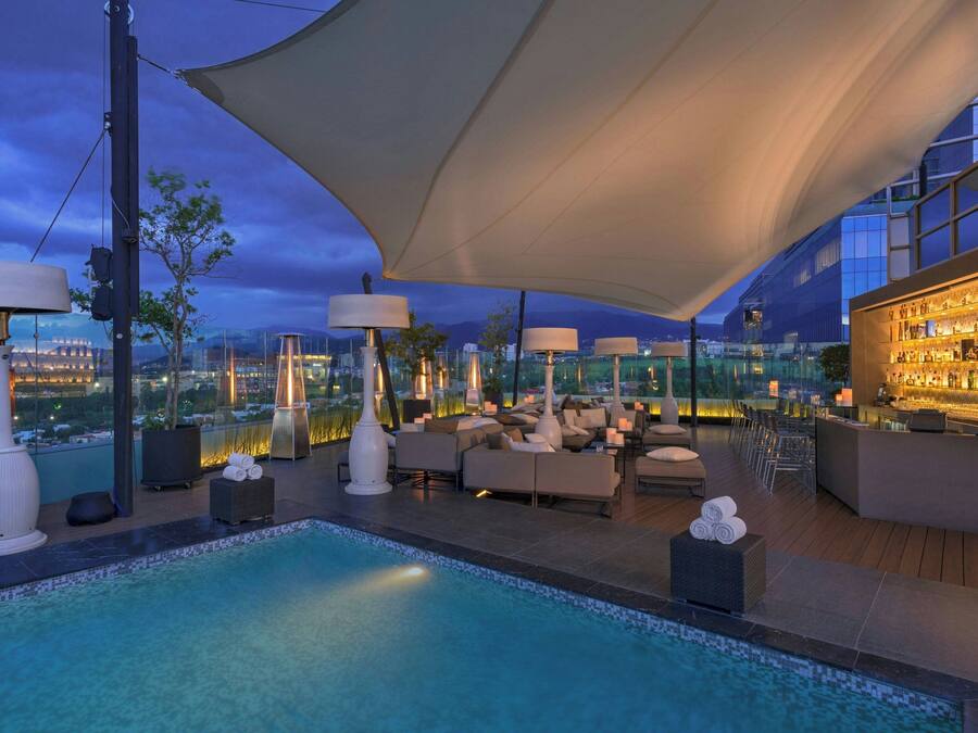 An outdoor pool area with lounge seating, a bar, and a city view at dusk.