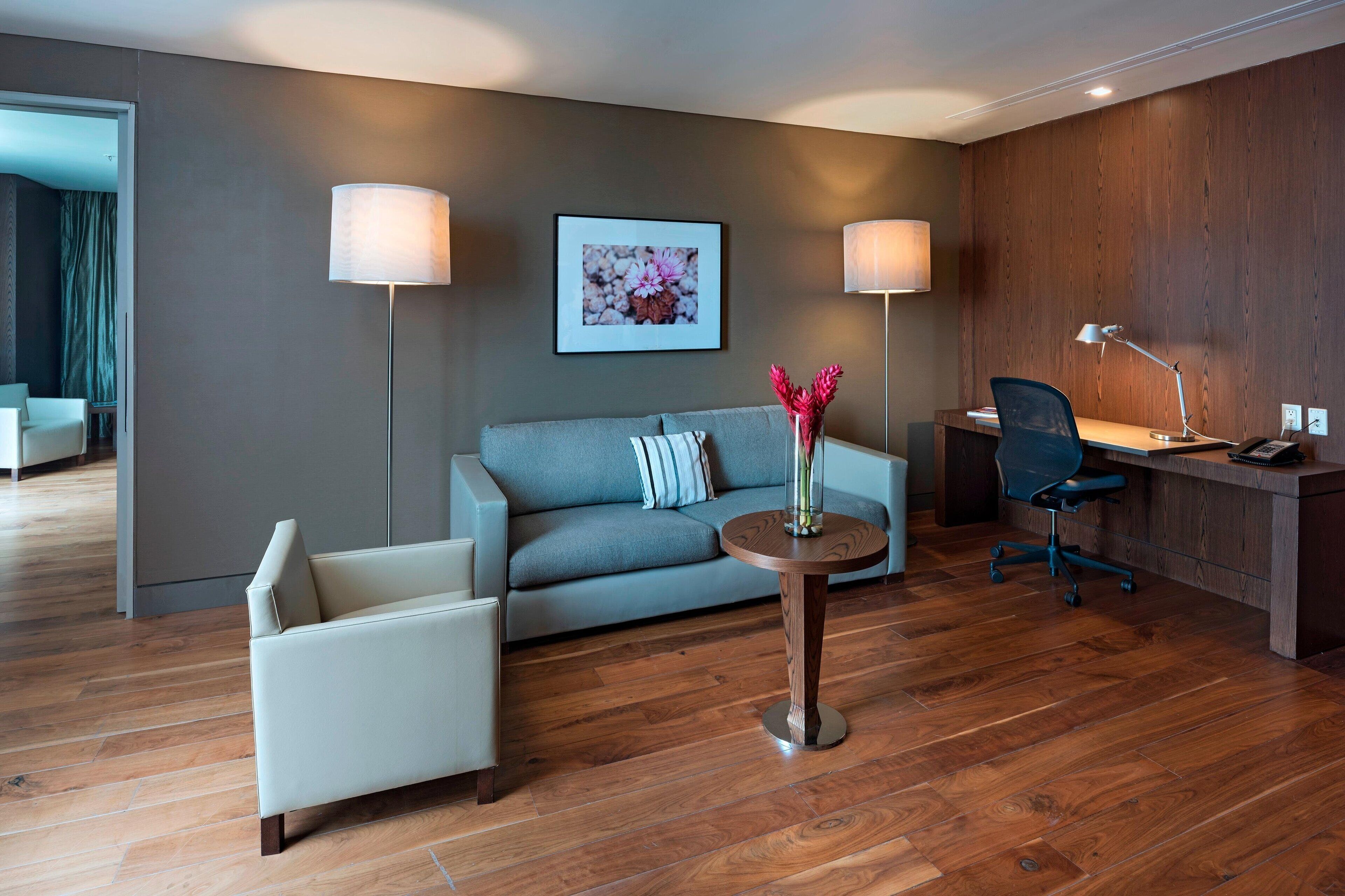 A modern hotel room with a grey sofa, a round wooden side table with a vase, a desk with a chair, and two lamps.