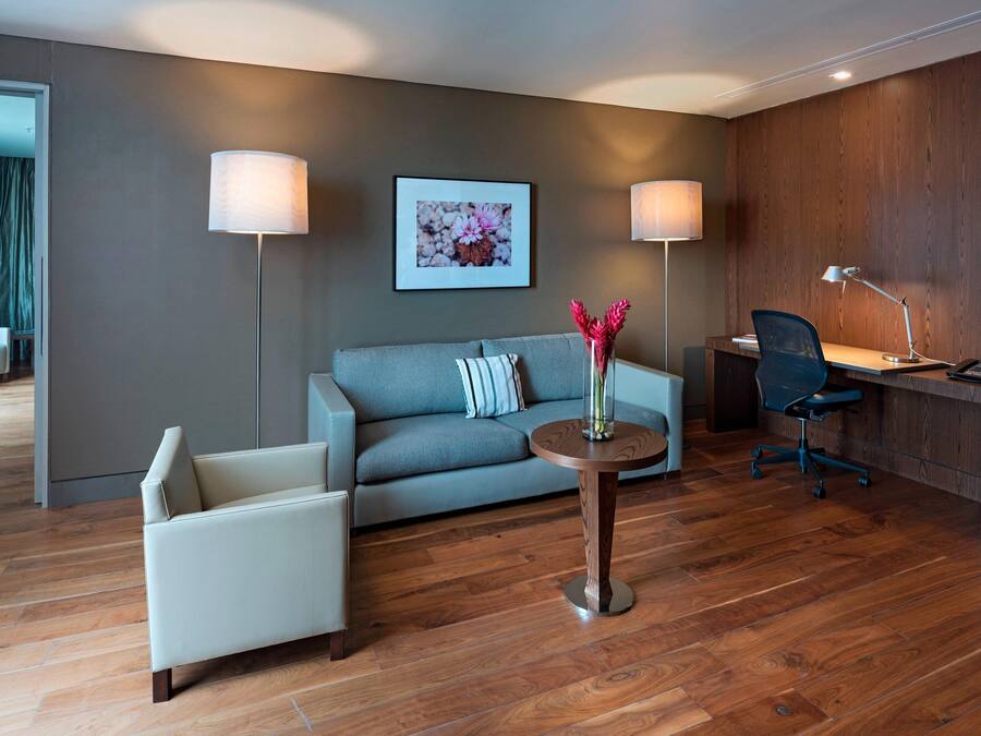A modern hotel room with a grey sofa, a round wooden side table with a vase, a desk with a chair, and two lamps.