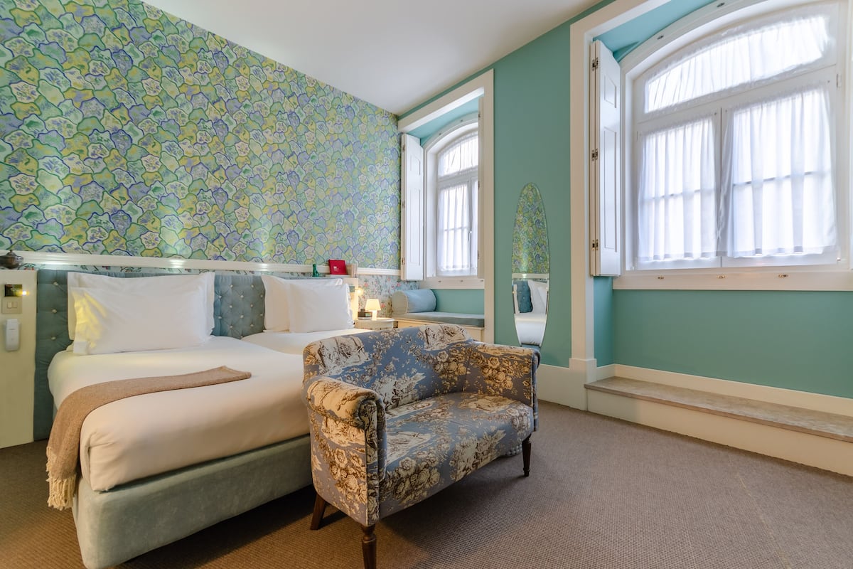 Een kamer met een bed, een bloemen fauteuil, een behang met patroon en een raam met gordijnen.
