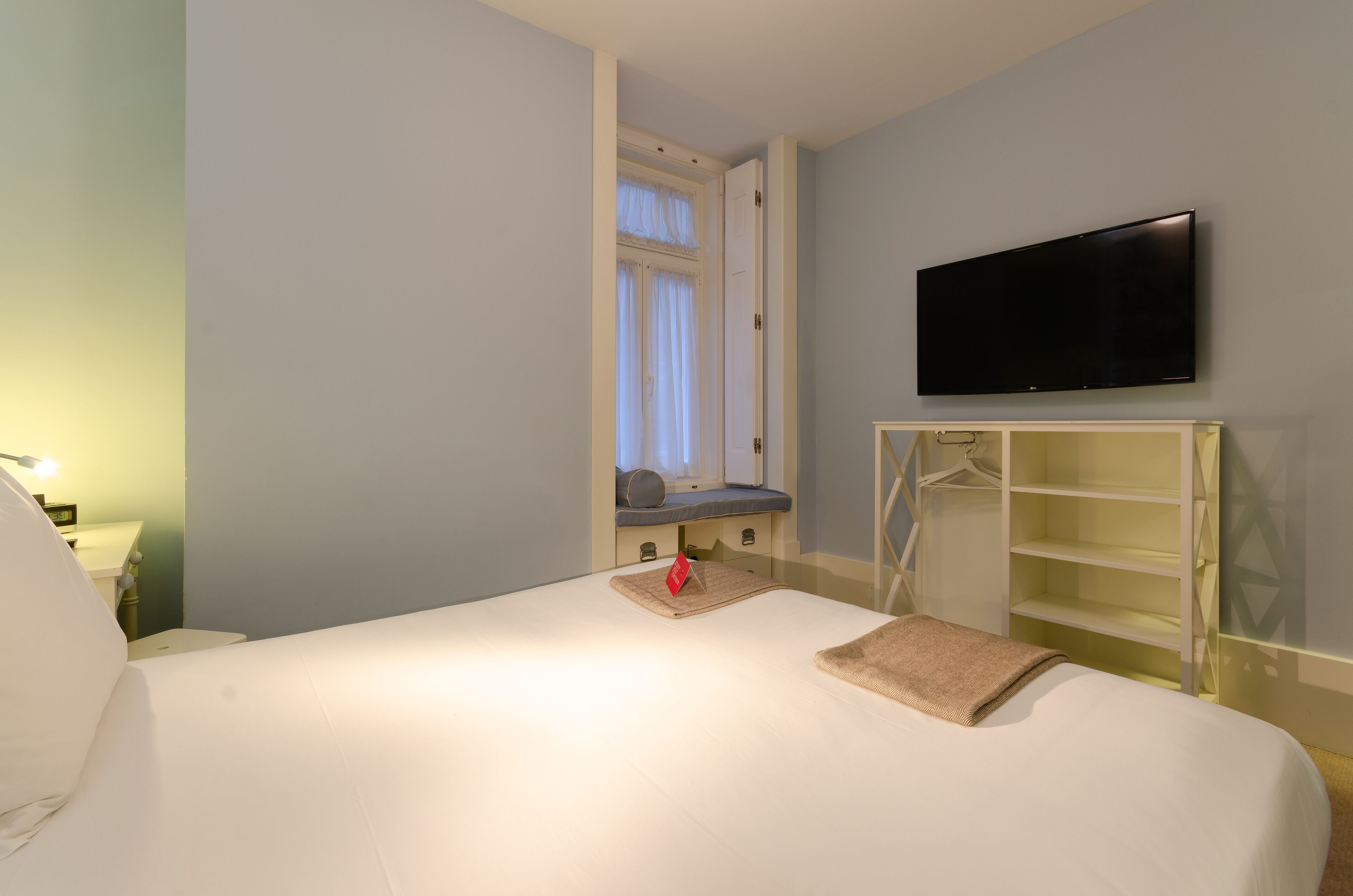 A hotel room with a bed, a TV mounted on the wall, a window with curtains, and a bedside table with a lamp.